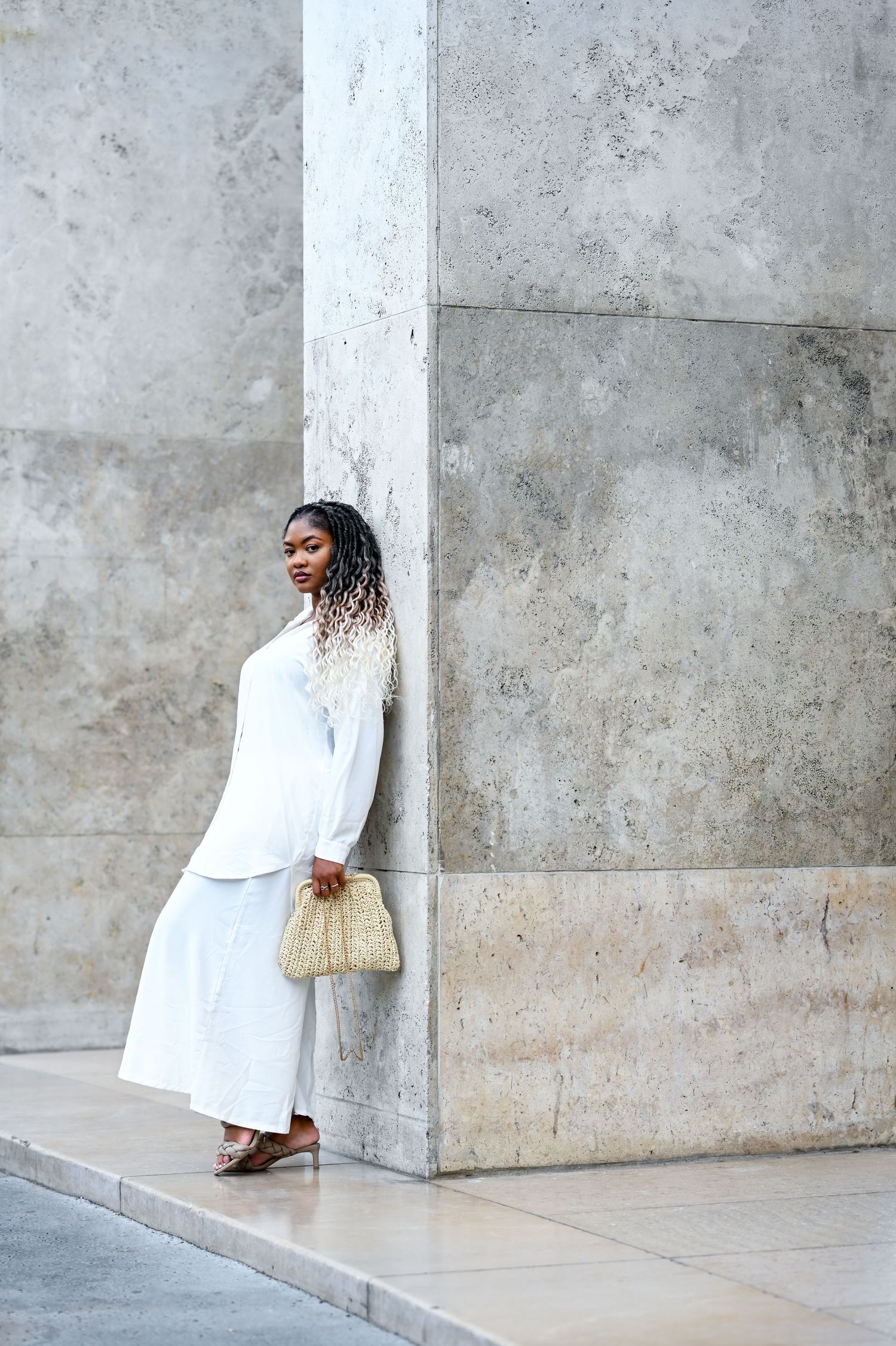 Model wearing flowing white dress stands between modern concrete pillars with a small beige handbag.