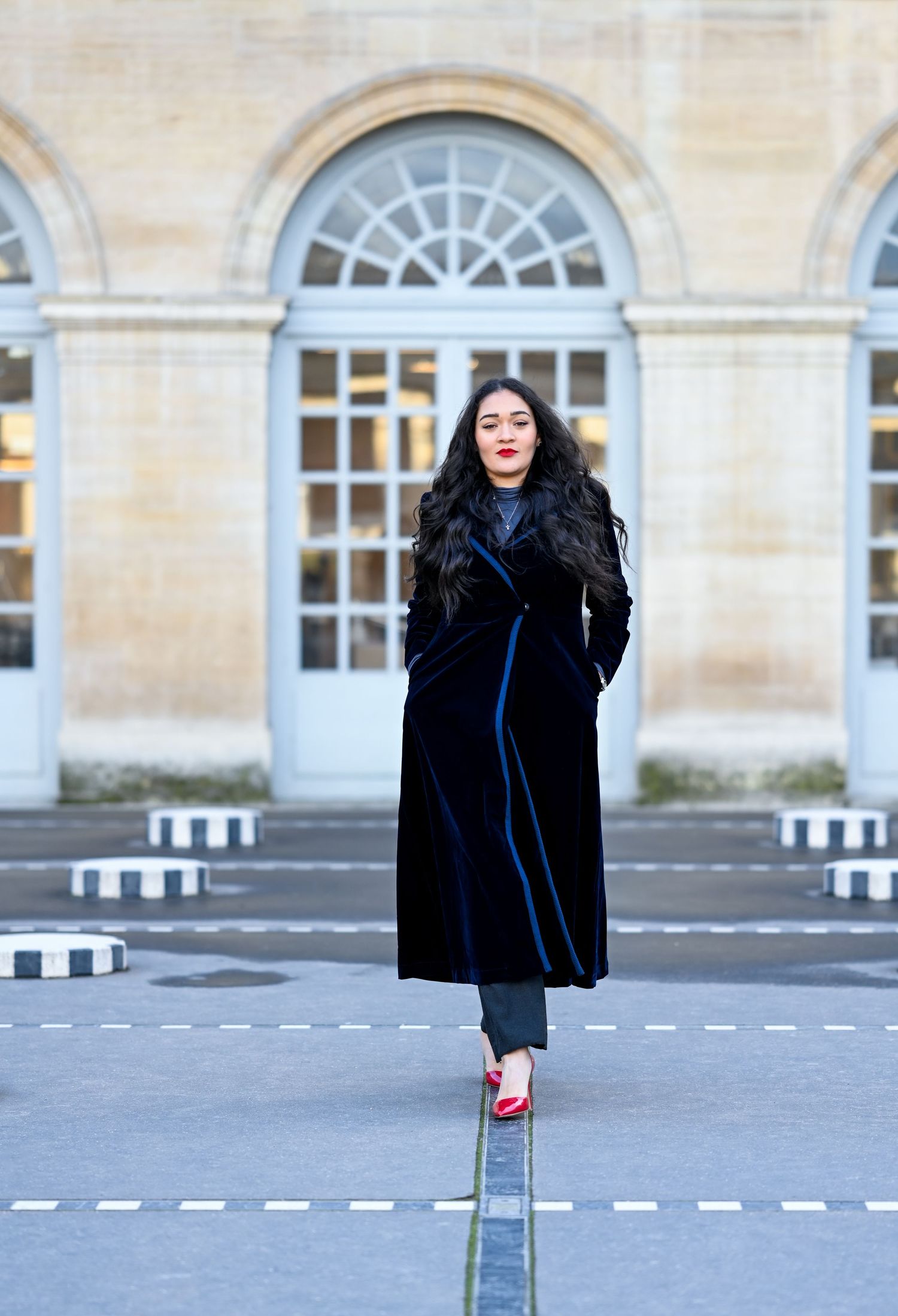 Fashionable woman in long black coat stands confidently against classical Parisian architecture.