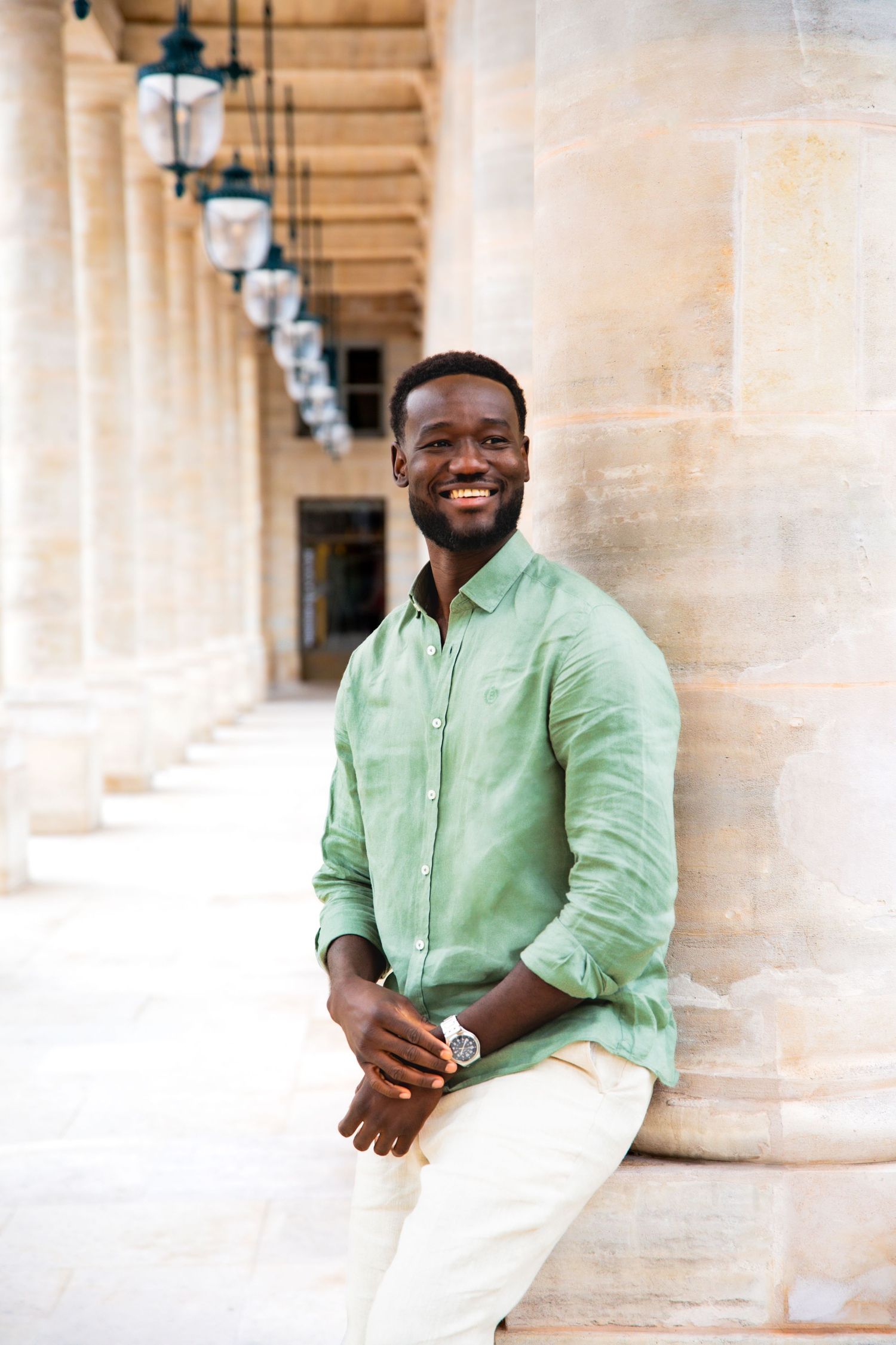 Smiling person in mint green linen shirt poses confidently against elegant stone columns and archways.