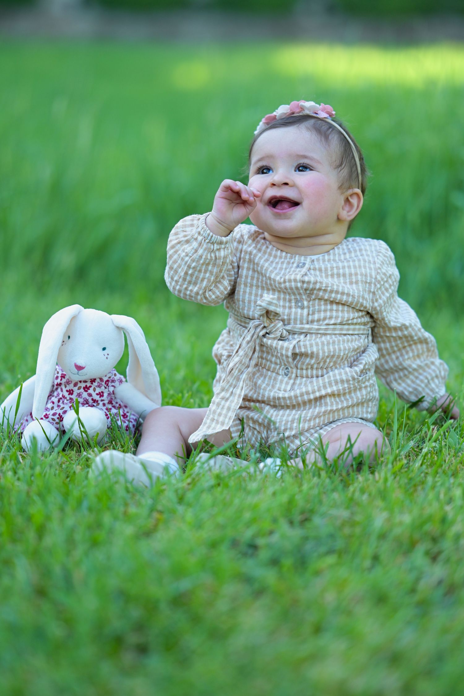 Baby in a checkered outfit lies on lush green grass with a plush bunny toy.