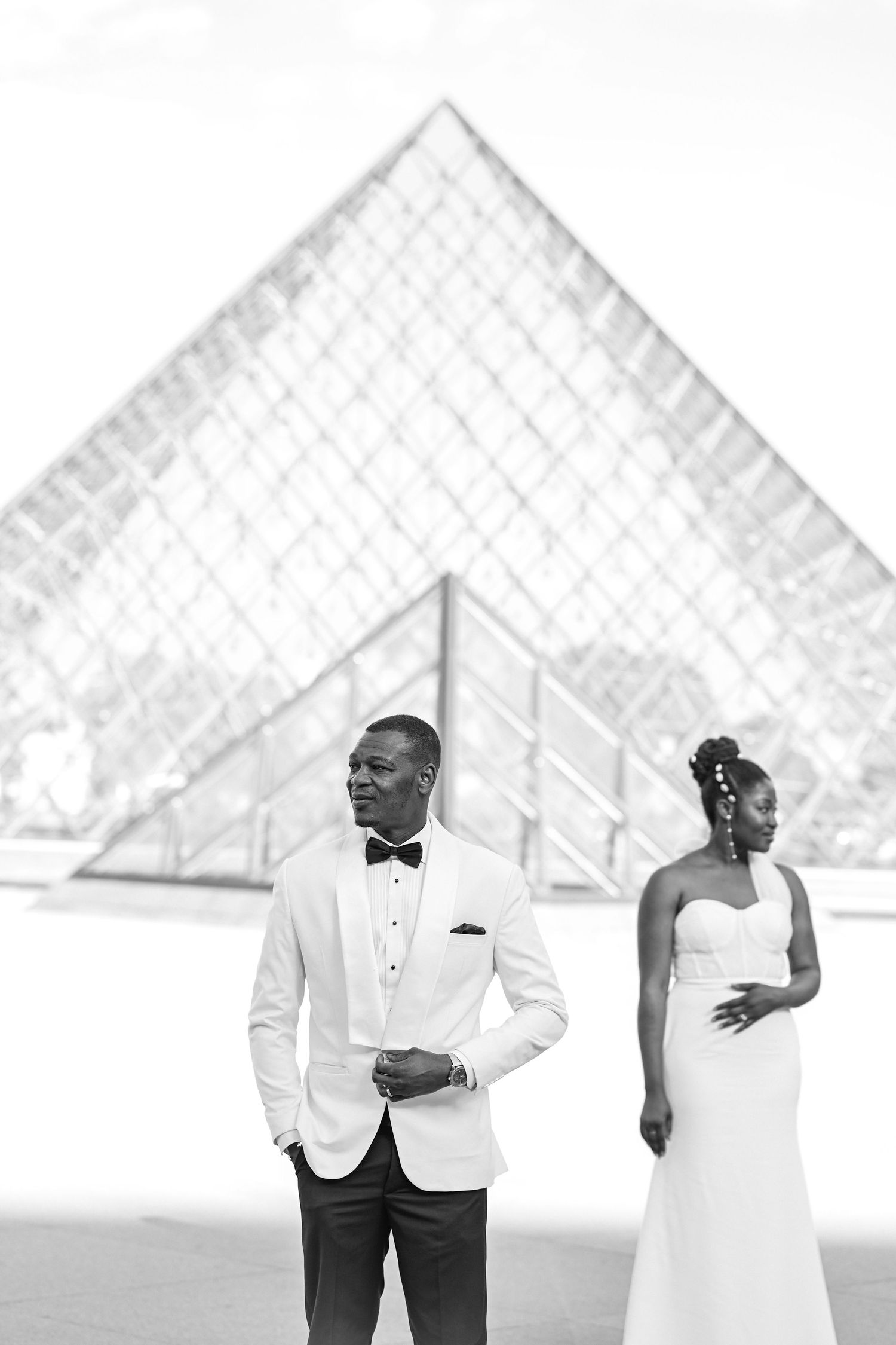 A couple in formal white attire poses in front of the iconic Louvre pyramid in Paris.