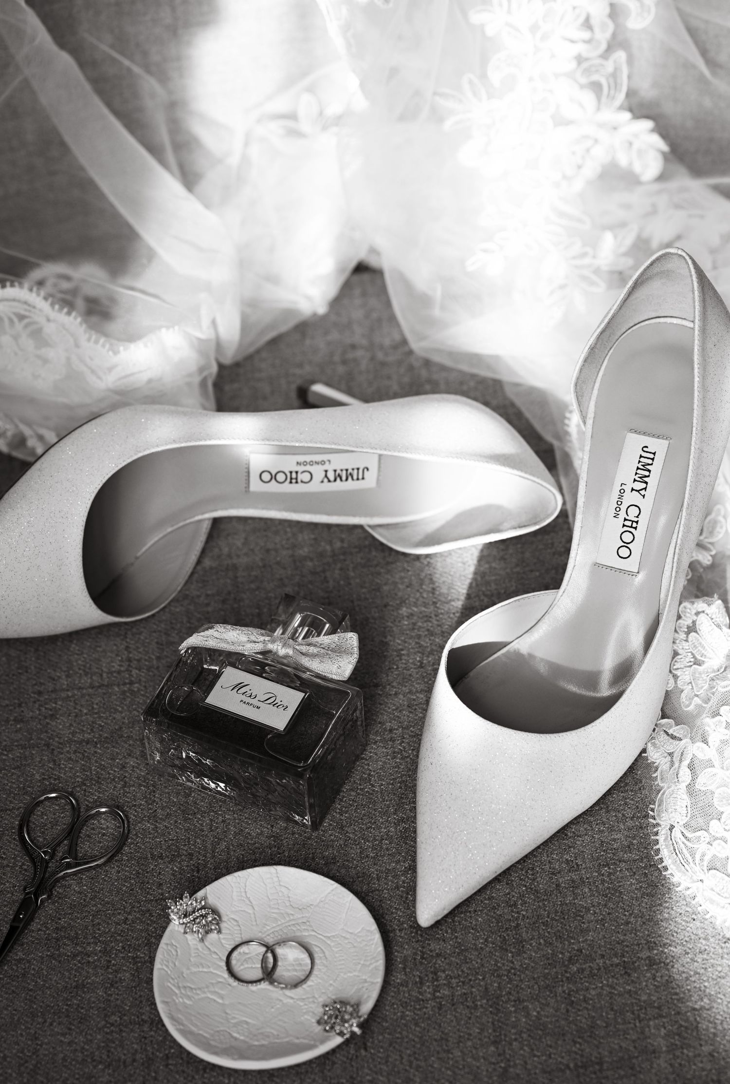 Wedding accessories including white Jimmy Choo heels, perfume, and wedding rings on lace fabric.