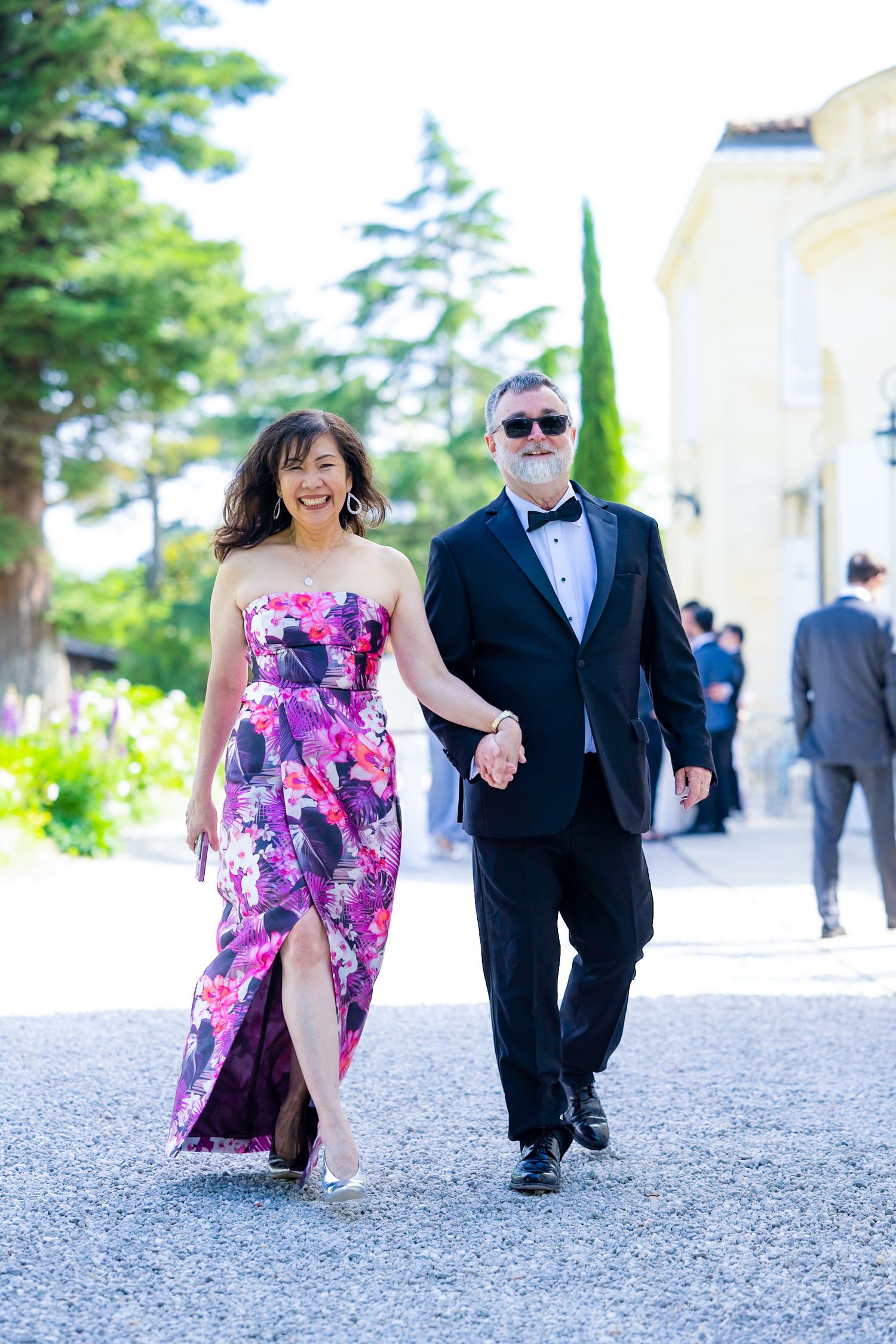 A couple walks arm in arm along a garden path, with the woman wearing a vibrant floral gown.