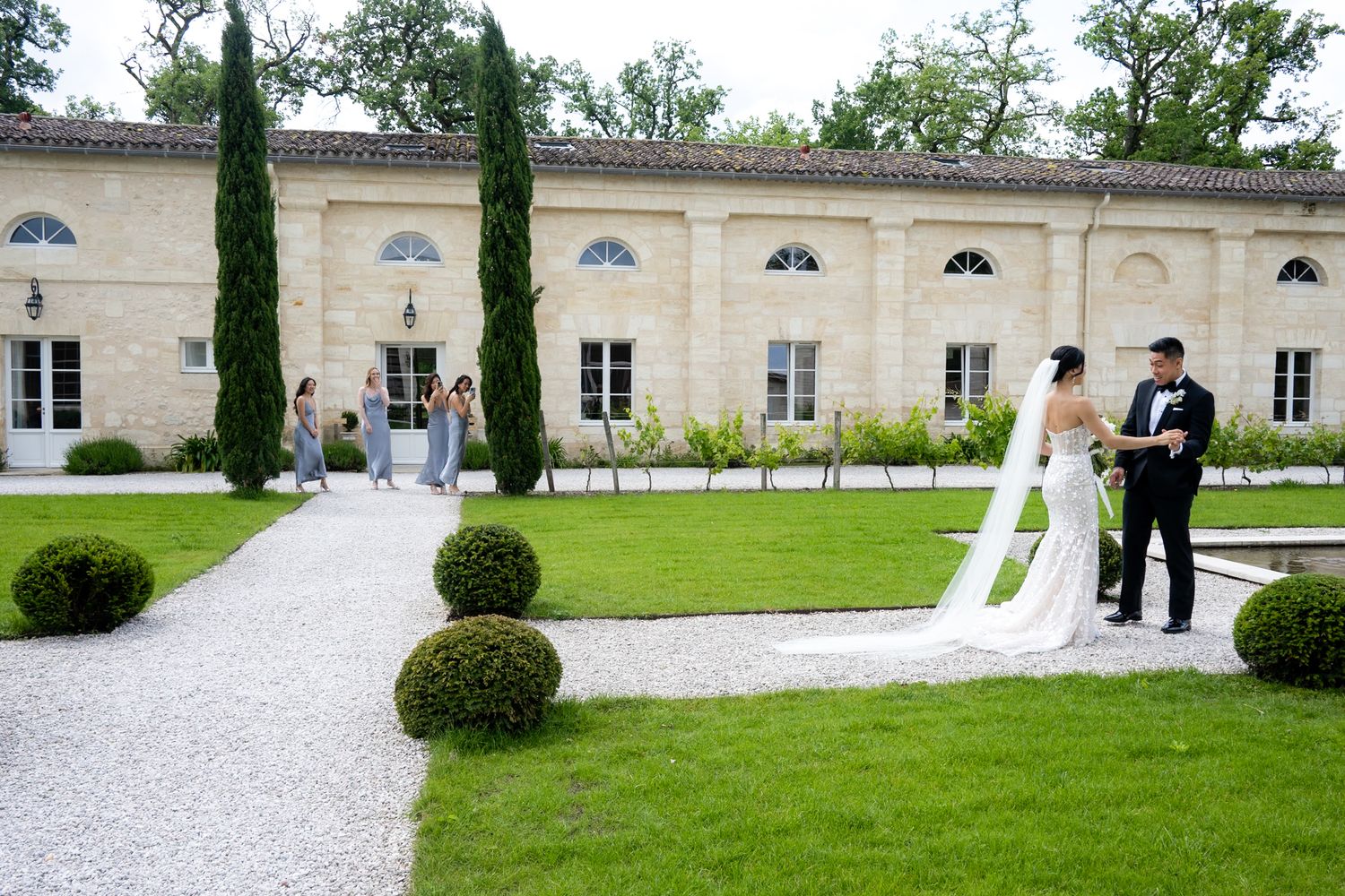 Wedding couple walking on gravel path outside historic stone mansion with manicured gardens.