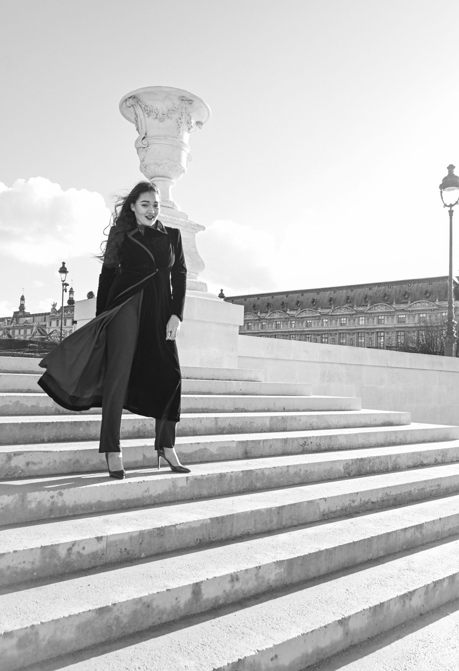 A figure in a long black coat descends elegant white marble steps in a classic black and white architectural scene.
