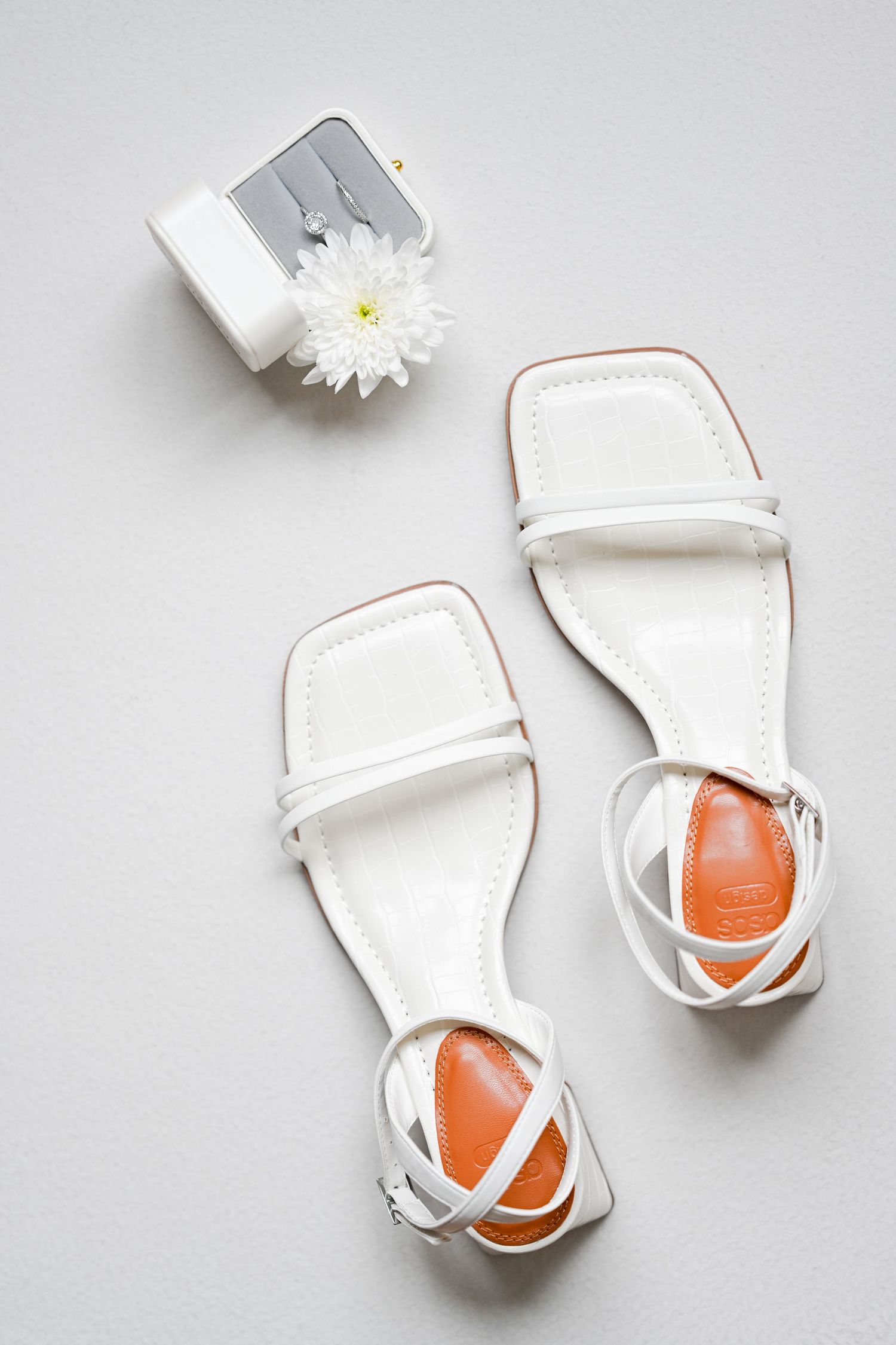 White strappy sandals and a daisy flower arranged on a light gray background.