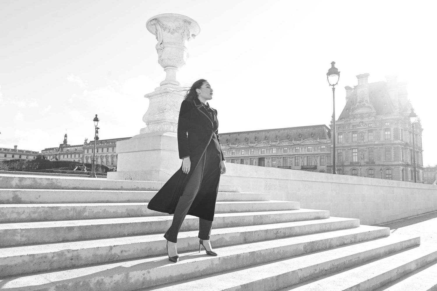 Woman in flowing black coat poses dramatically on white stone steps in Paris.