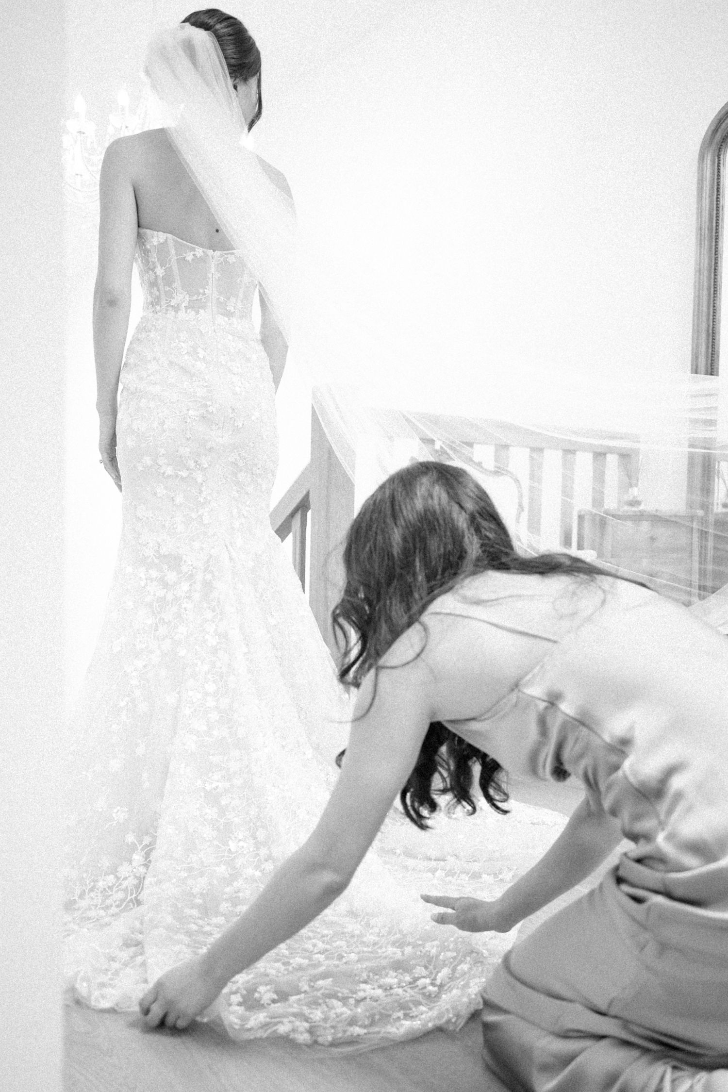 Bridesmaid adjusting the train of a lace wedding dress on a bride wearing a veil, black and white photo.