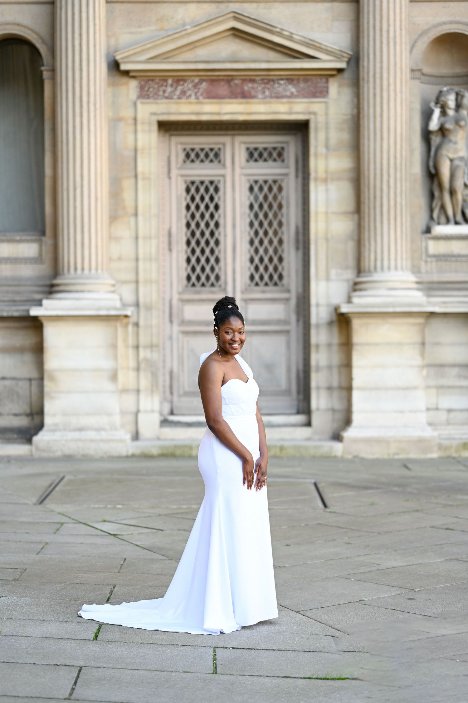A person in a white dress stands elegantly against a classical architectural backdrop.