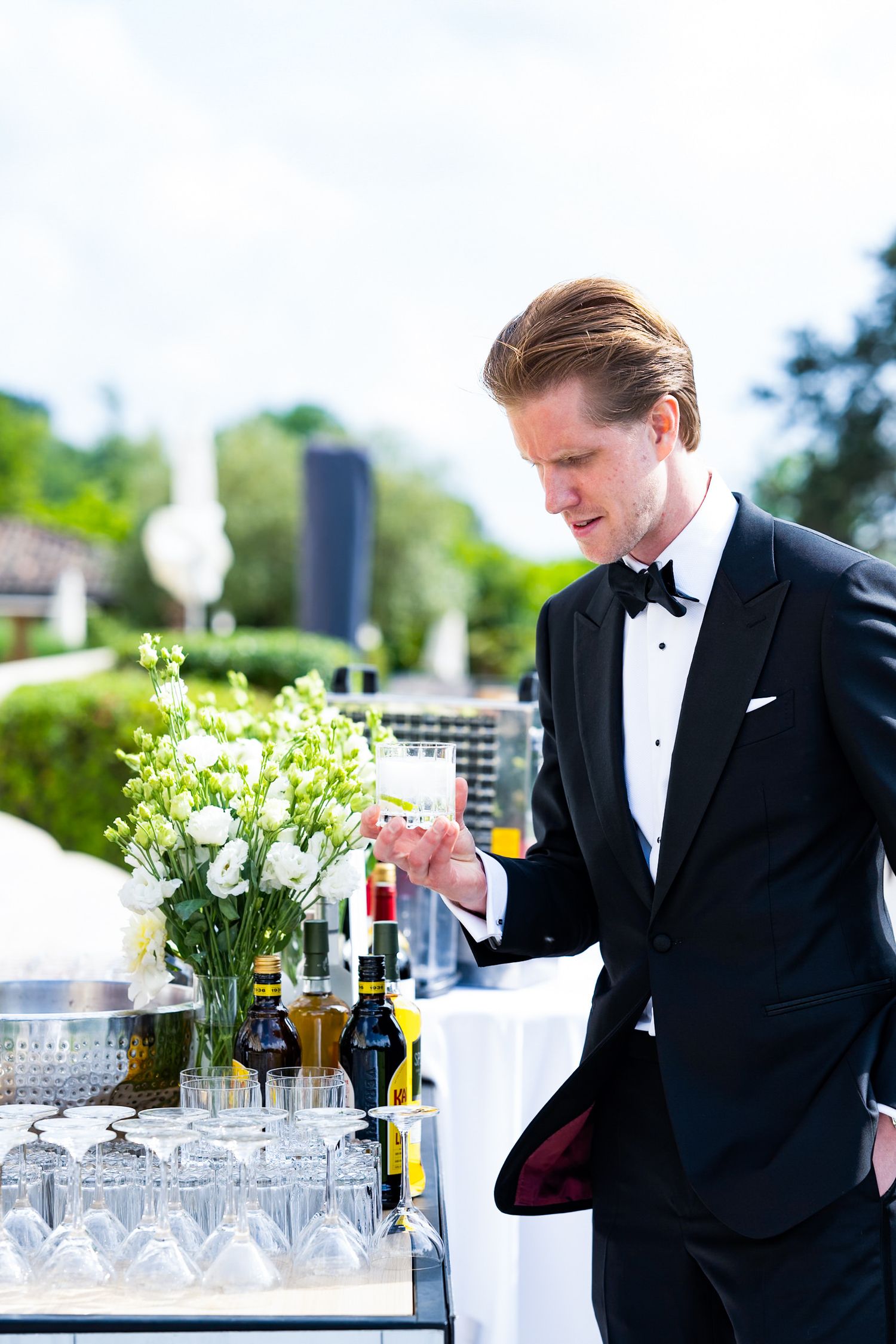 A guest in a black tuxedo arranges drinks at an elegant outdoor bar setup with crystal glassware.