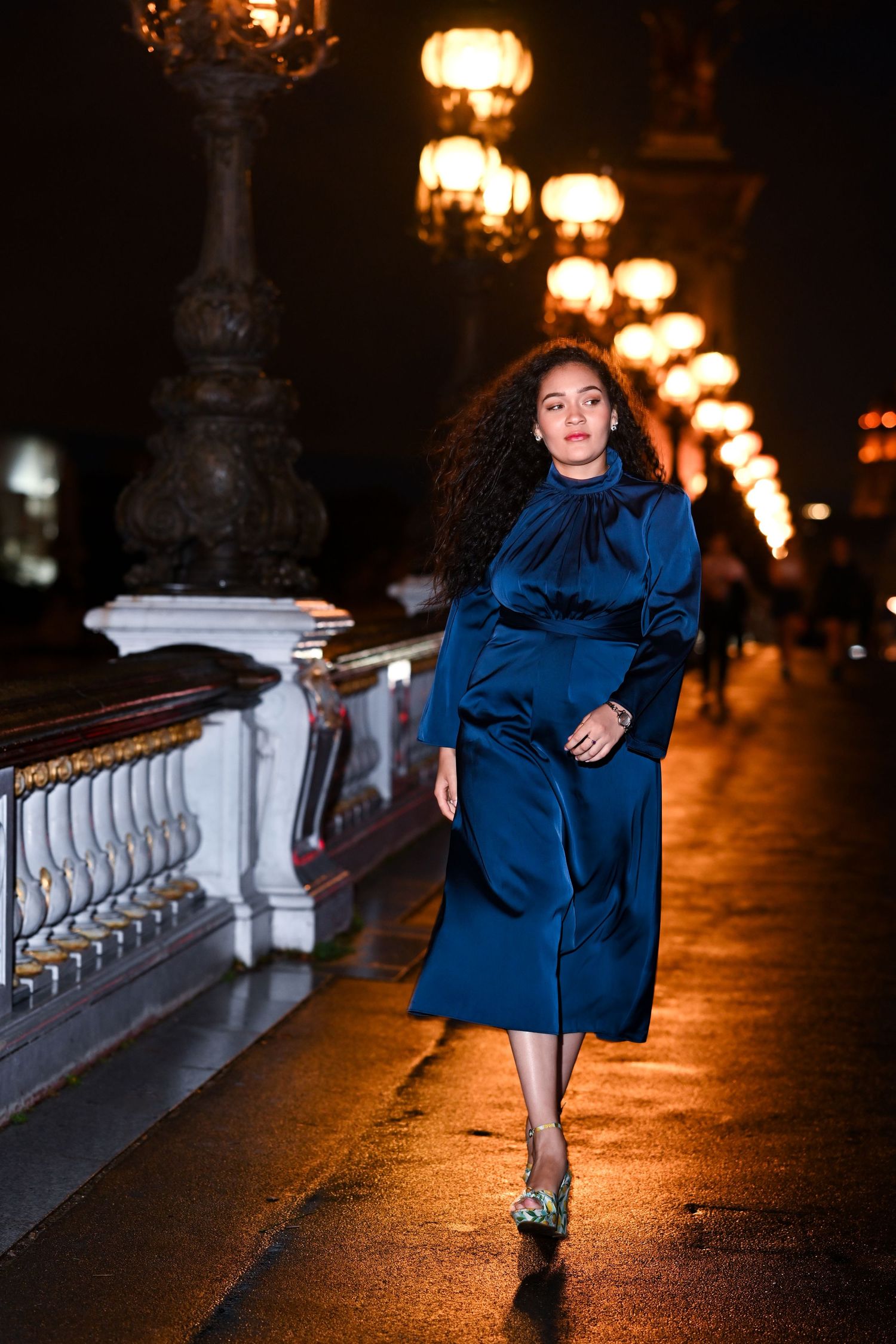 Person in blue dress walking along illuminated bridge at night with vintage street lamps.