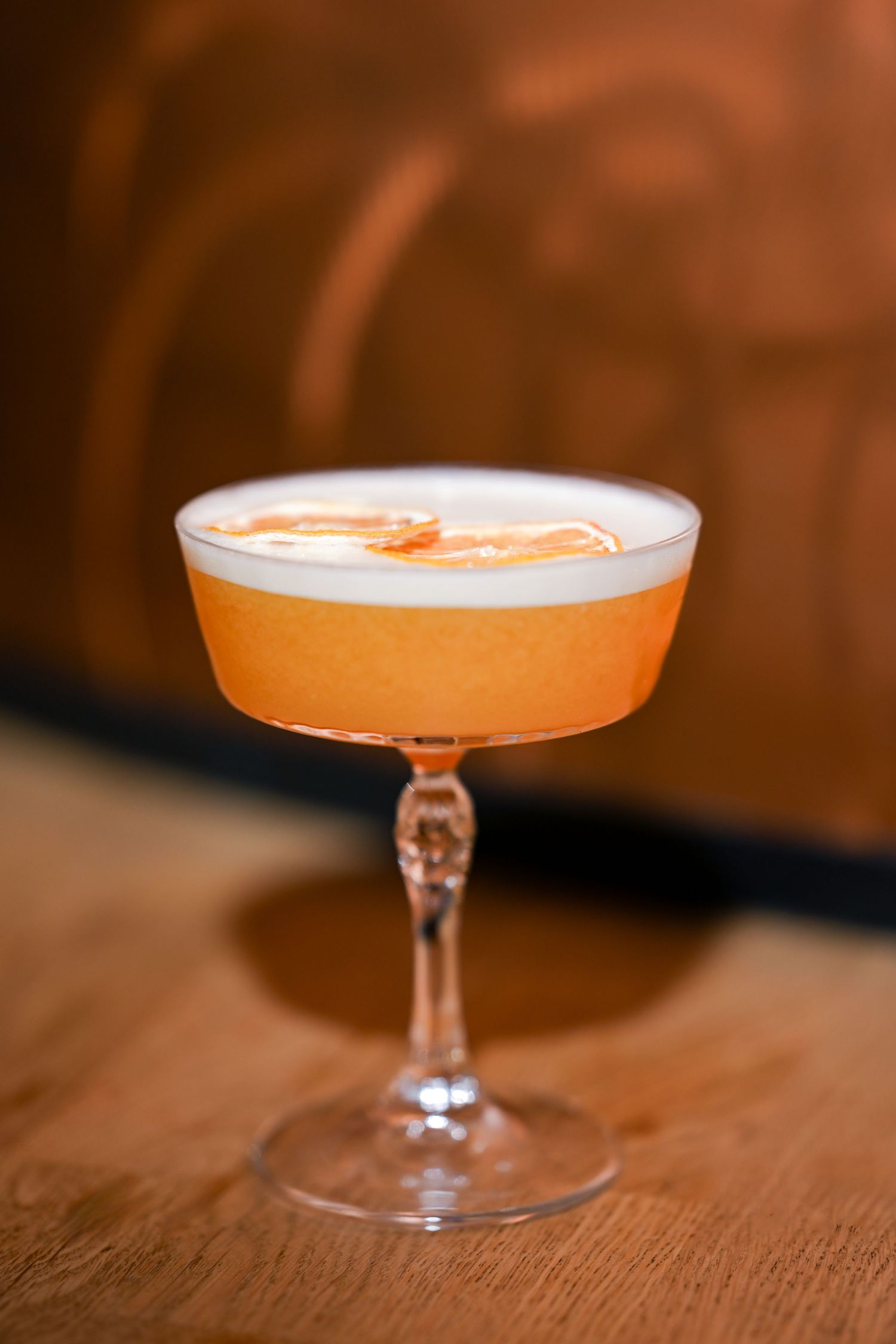 Orange craft cocktail with white foam served in a coupe glass on wooden surface.