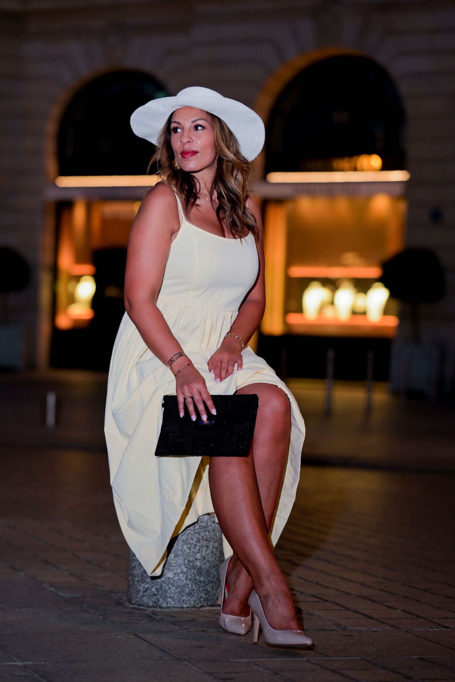 Elegant pose in white sundress and wide-brimmed hat against warm evening backdrop.