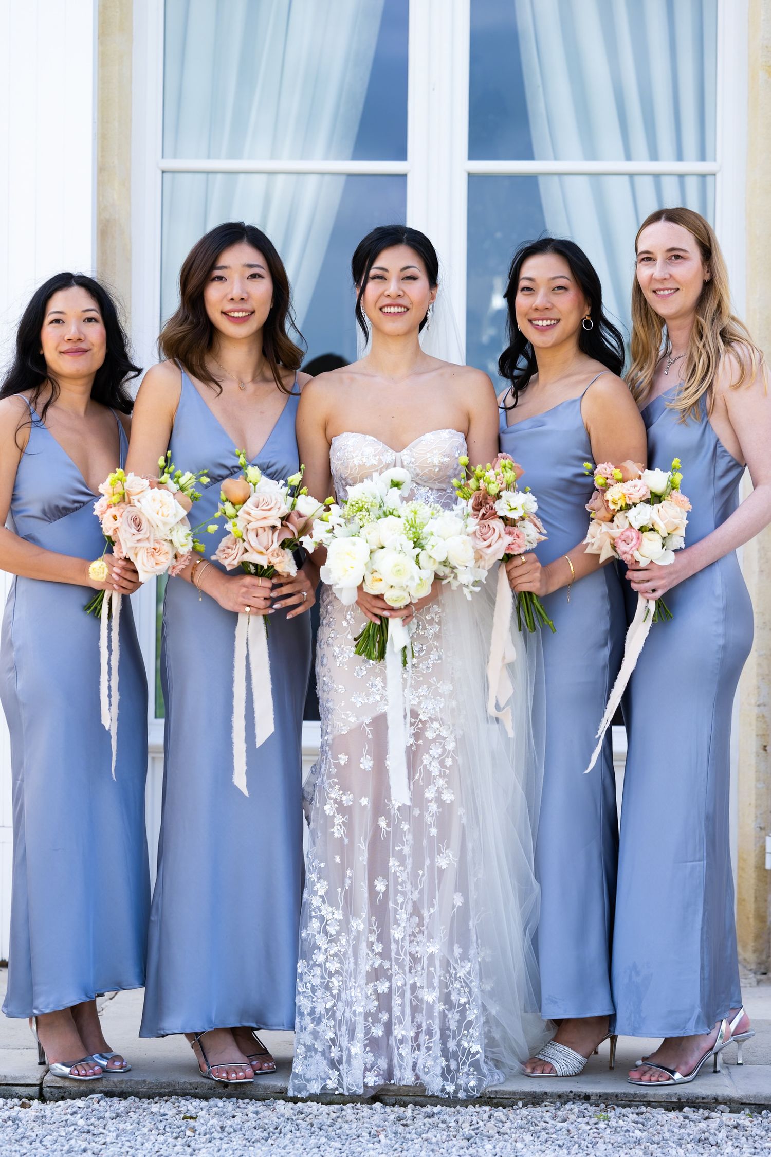 Bridesmaids in periwinkle blue dresses pose with bouquets alongside the bride in a lace gown.