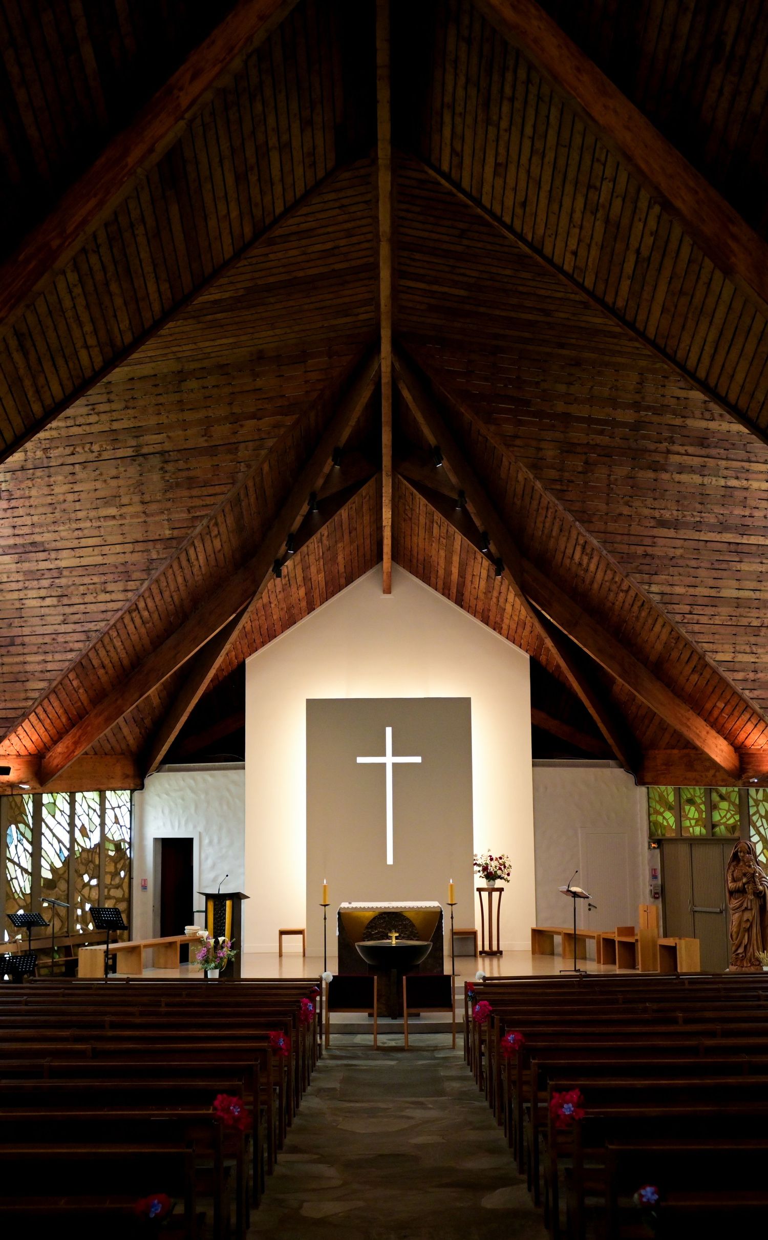 Interior of rustic church with exposed wooden beams and illuminated white cross at the altar.
