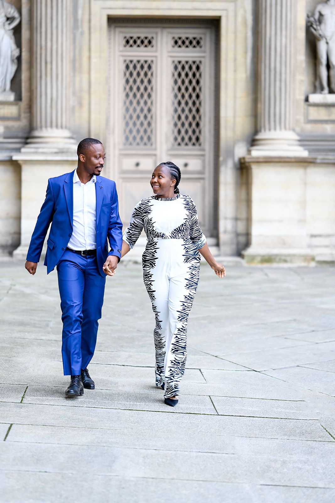 A couple in blue suit and patterned outfit dance and play on a stone courtyard.