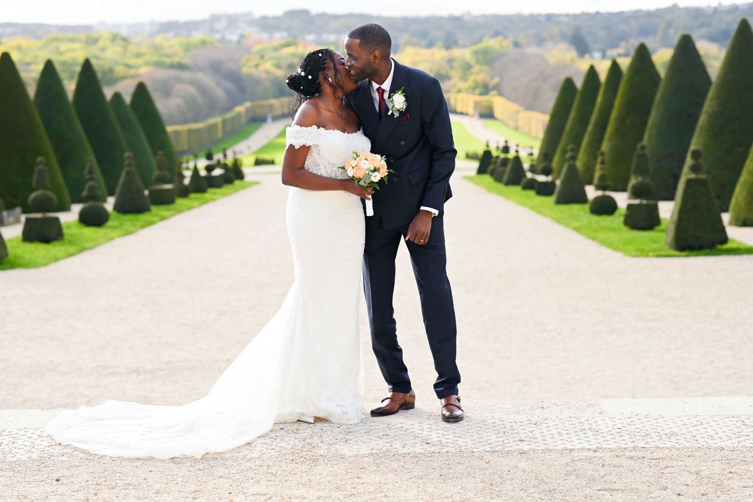 Elegant wedding couple strolls down a formal garden path with symmetrical evergreen topiaries.