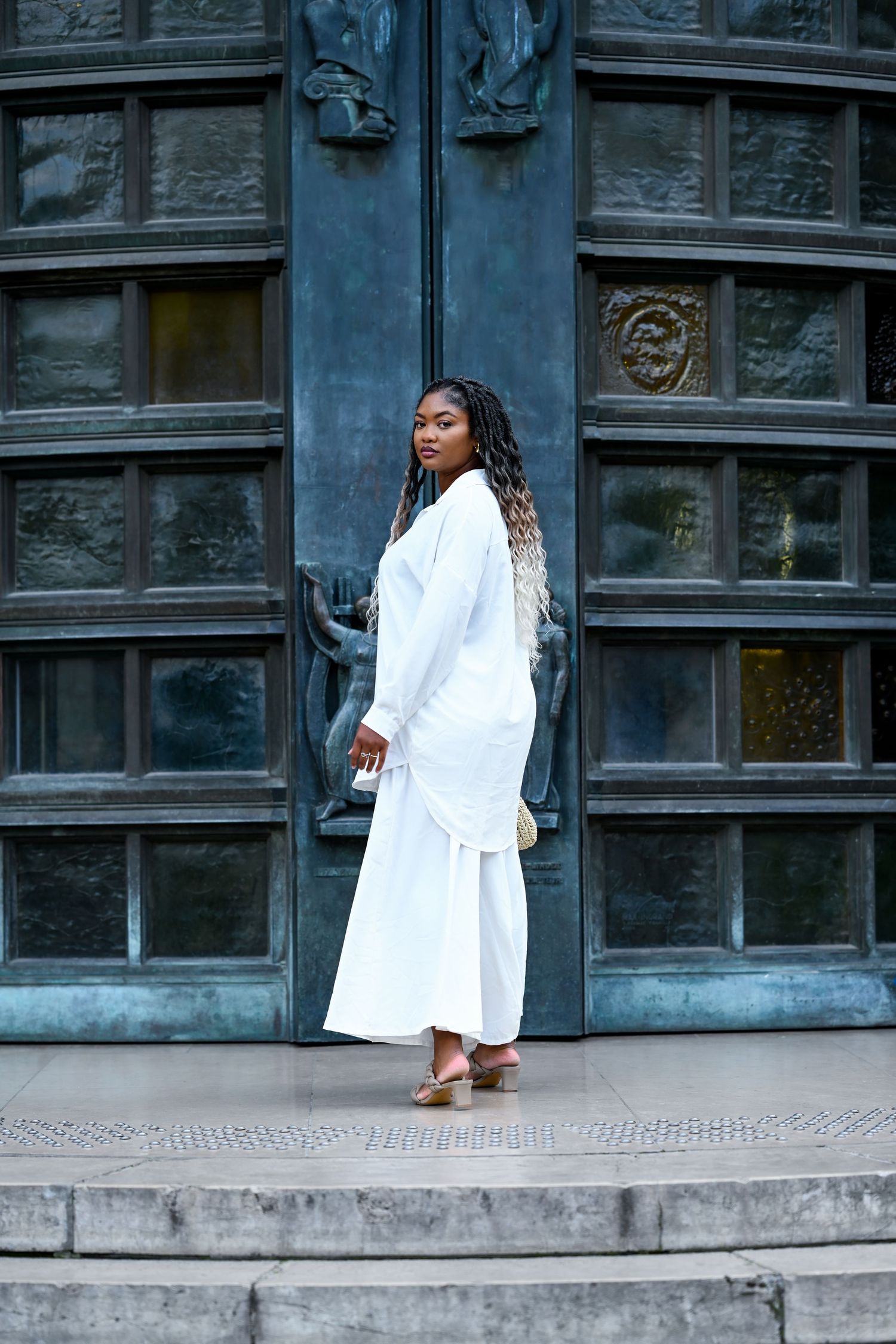 Person in white ensemble poses against dark architectural backdrop with ornate details.