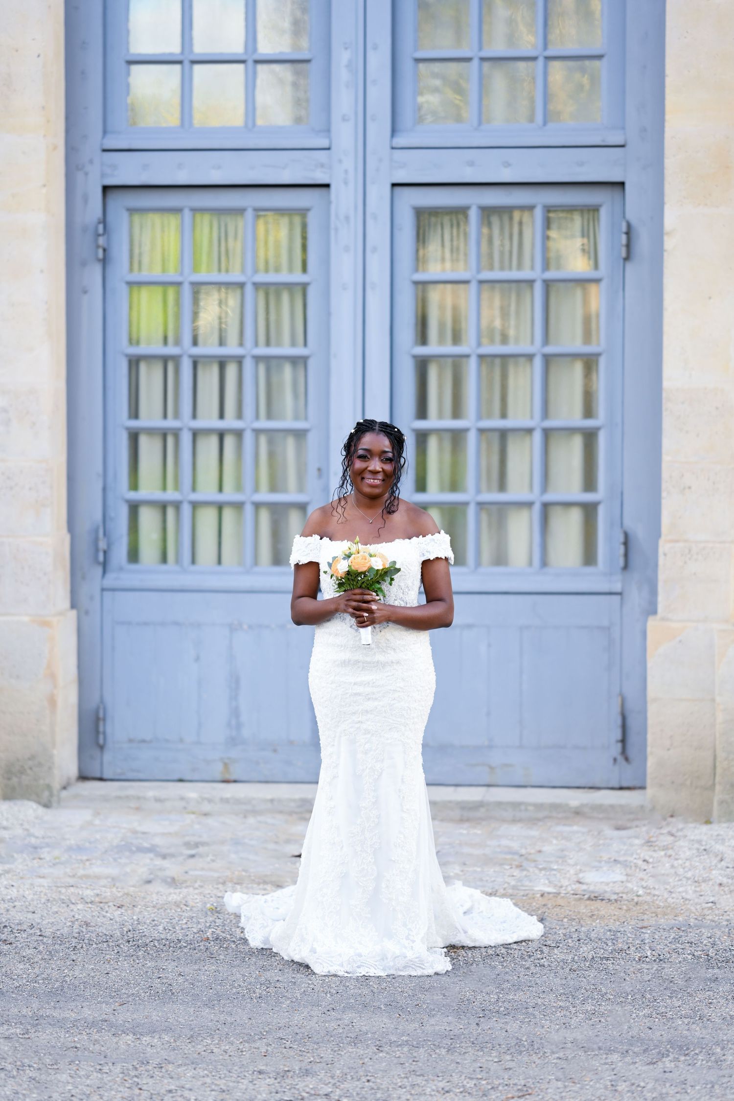 Elegant bride in off-shoulder lace gown posing in front of blue French doors.