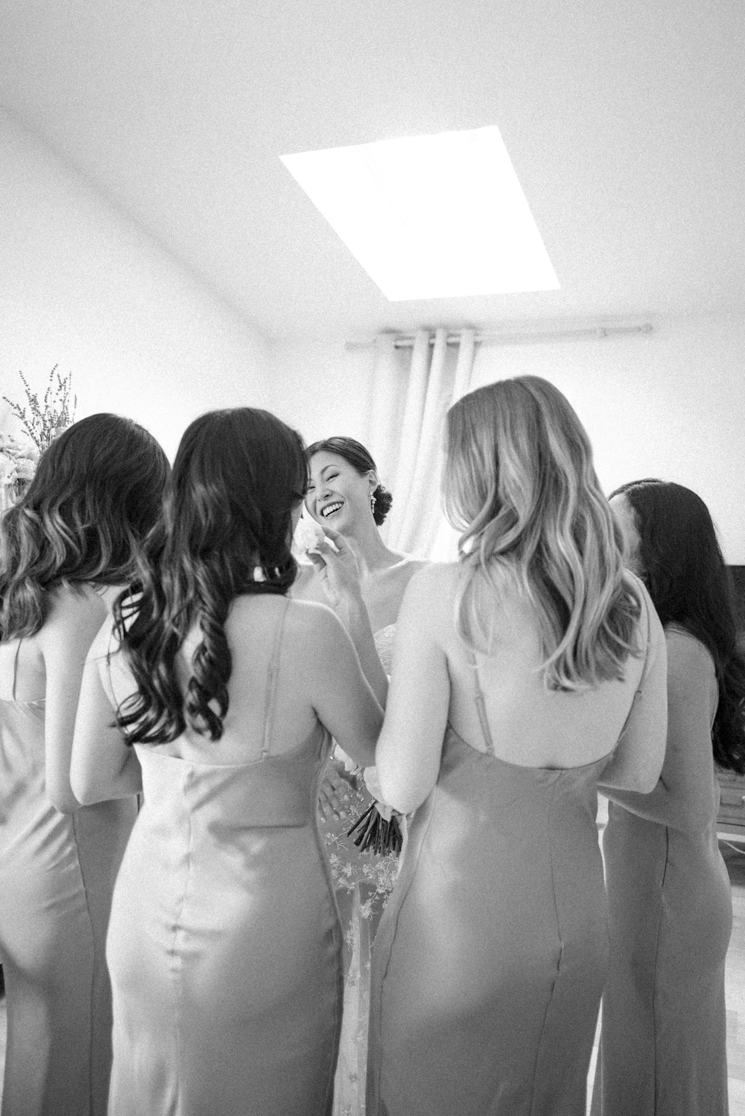 Black and white photo of a bride laughing with bridesmaids in satin dresses during wedding preparations.