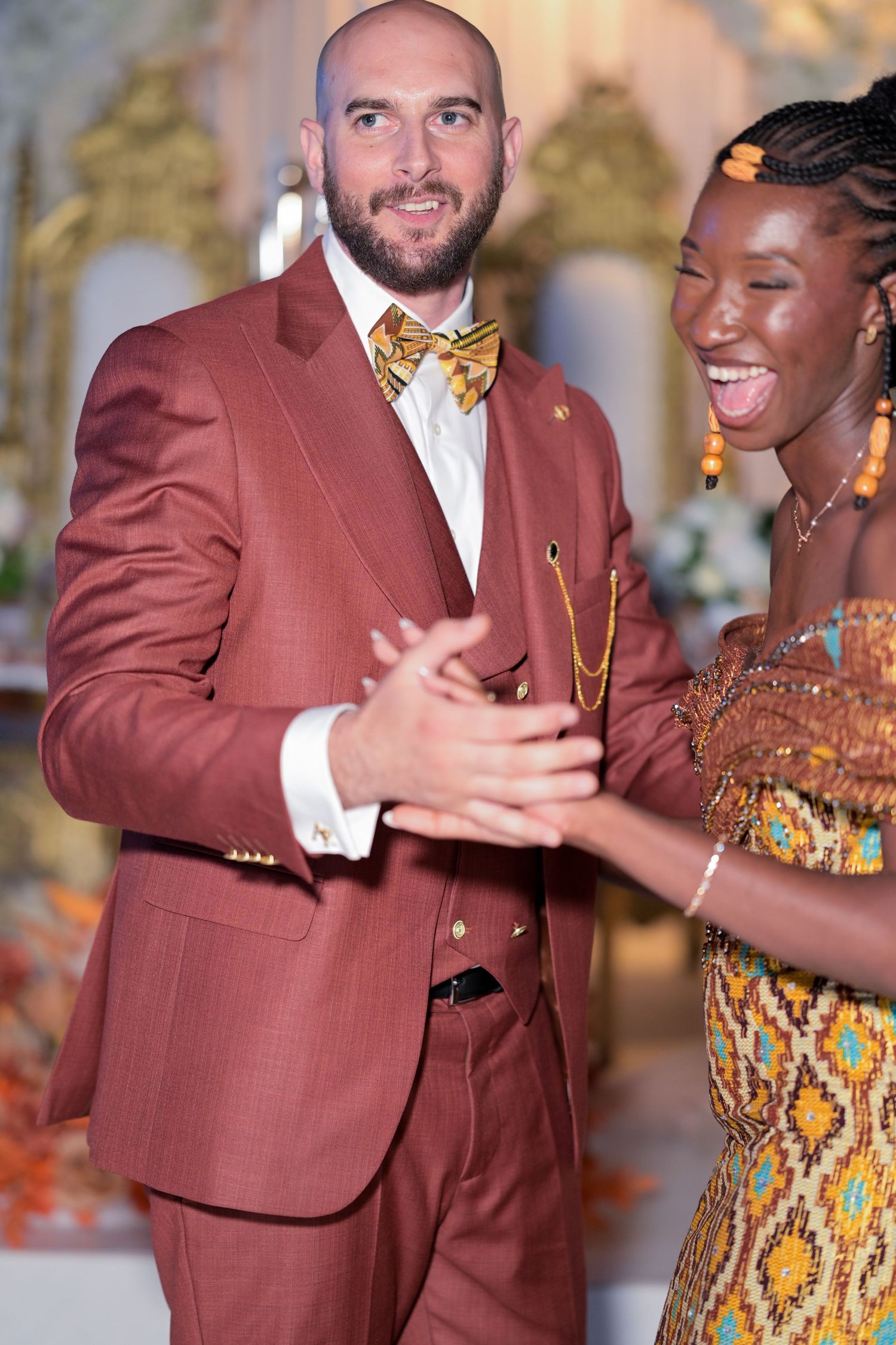 Man in rust-colored suit dancing with partner in traditional African print dress at formal event.