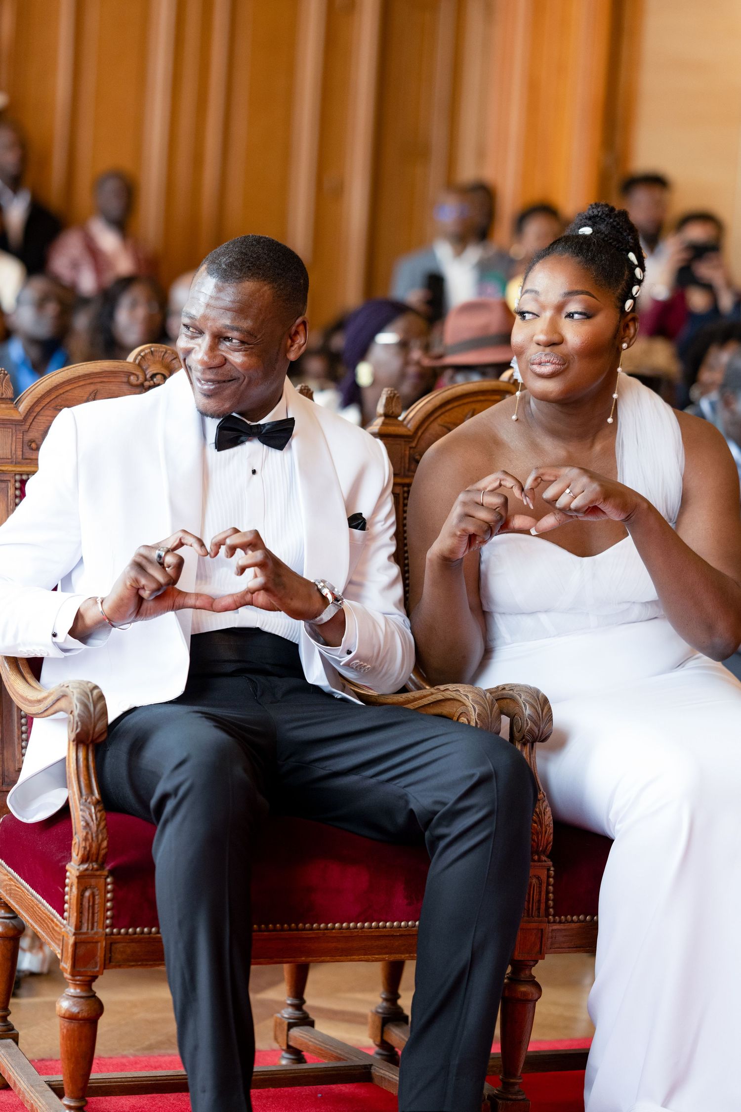 A couple in formal wedding attire making heart gestures while seated.