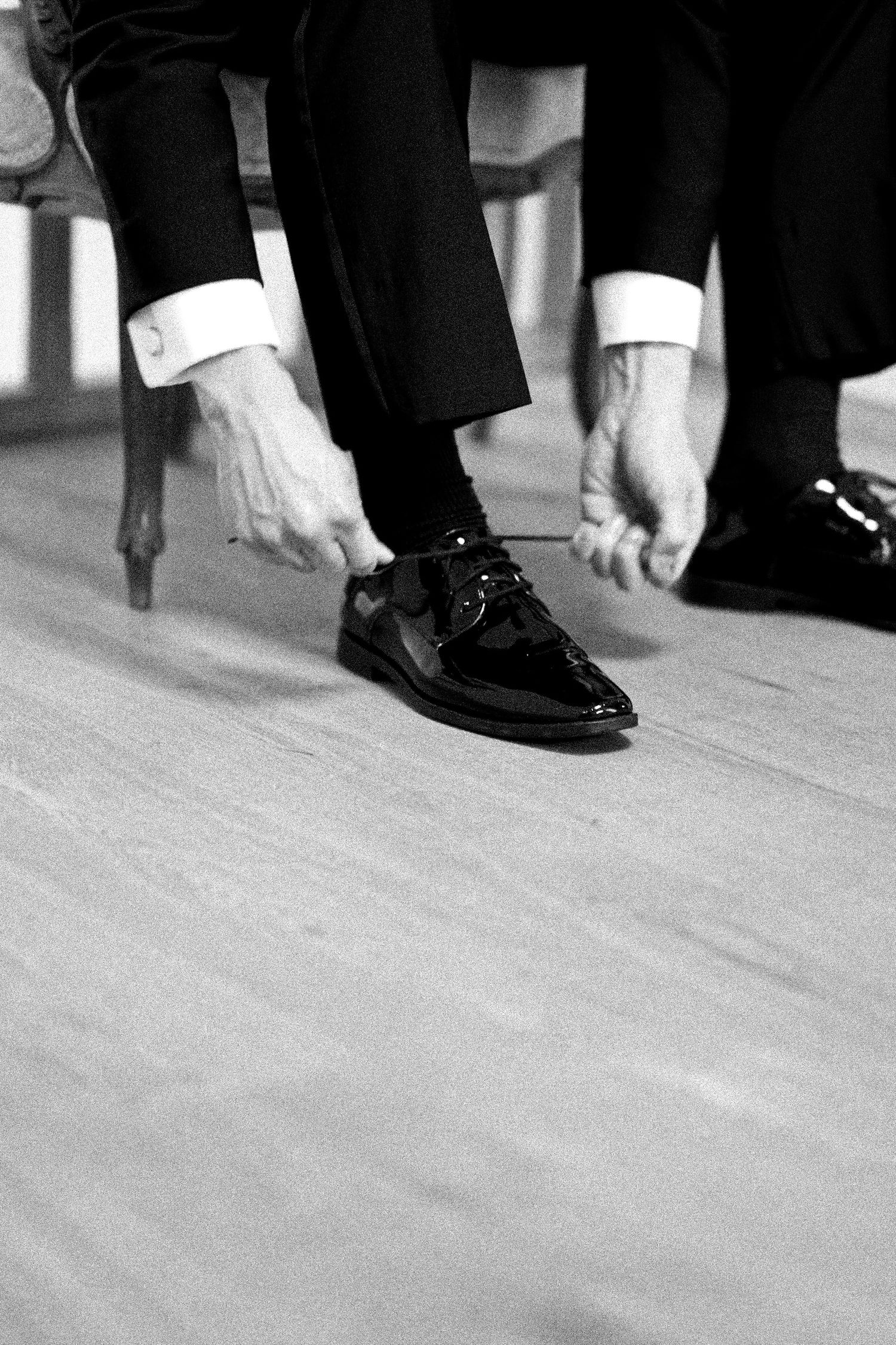 Black and white photo of hands adjusting formal dress shoes and cuffs on hardwood floor.
