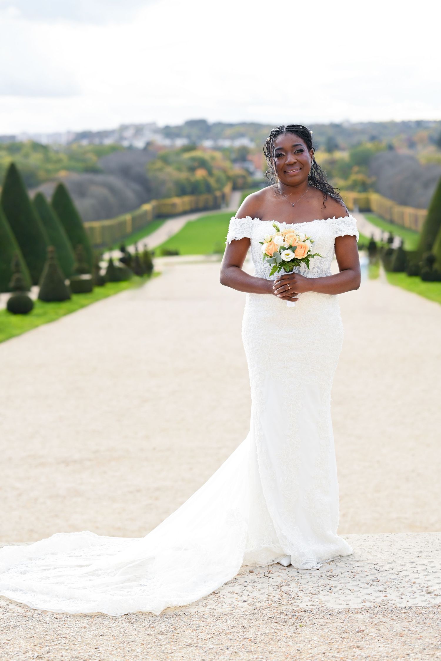 Bride in flowing white gown poses elegantly in a formal garden with manicured topiary backdrop.