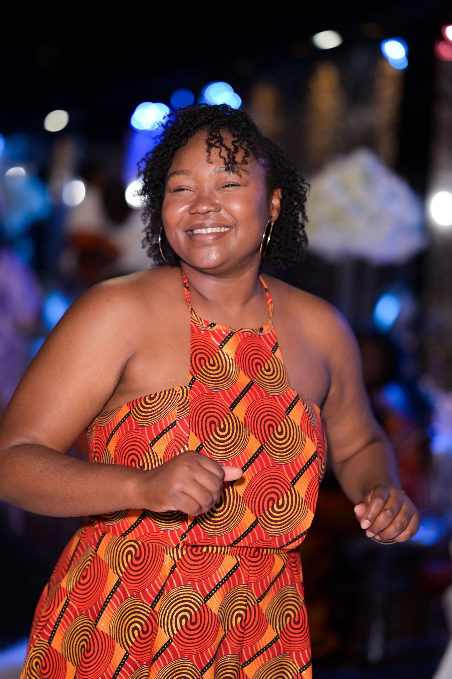 A person wearing a vibrant red and orange patterned dress dances enthusiastically at a celebration.