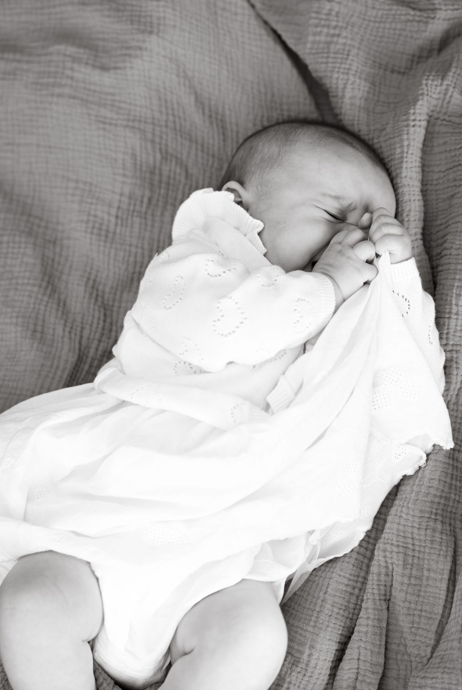 A sleeping baby in white clothing rests peacefully on grey bedding.