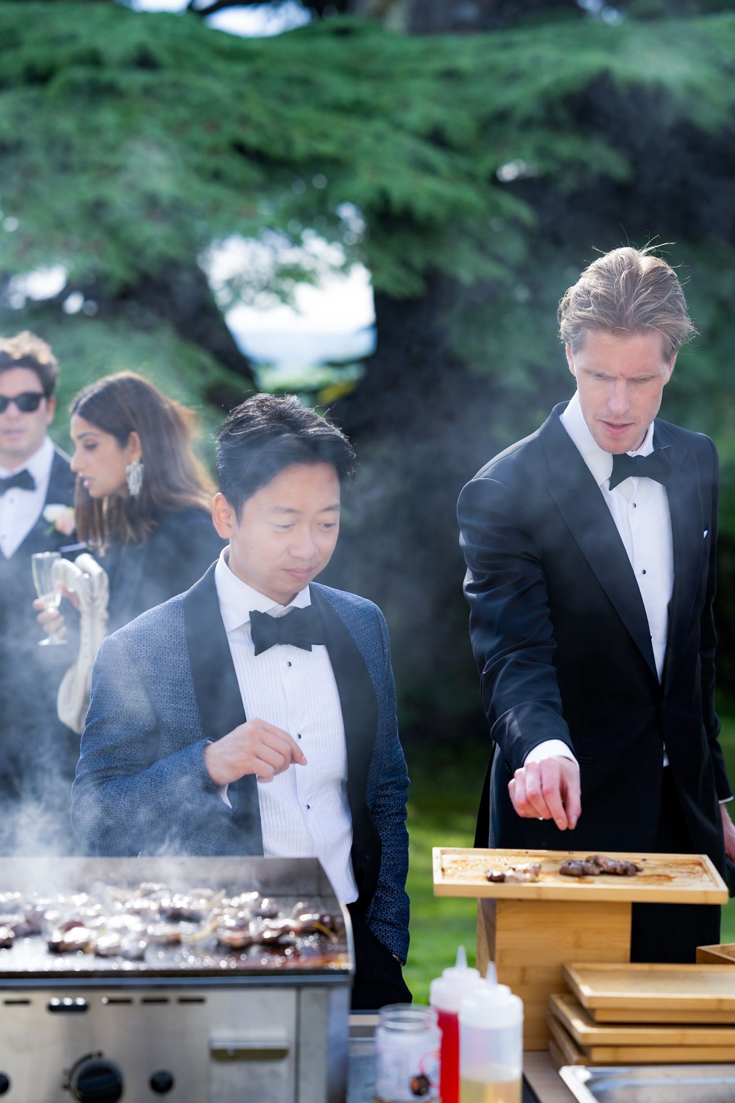 Two guests in tuxedos prepare food at an outdoor grill station during an event.
