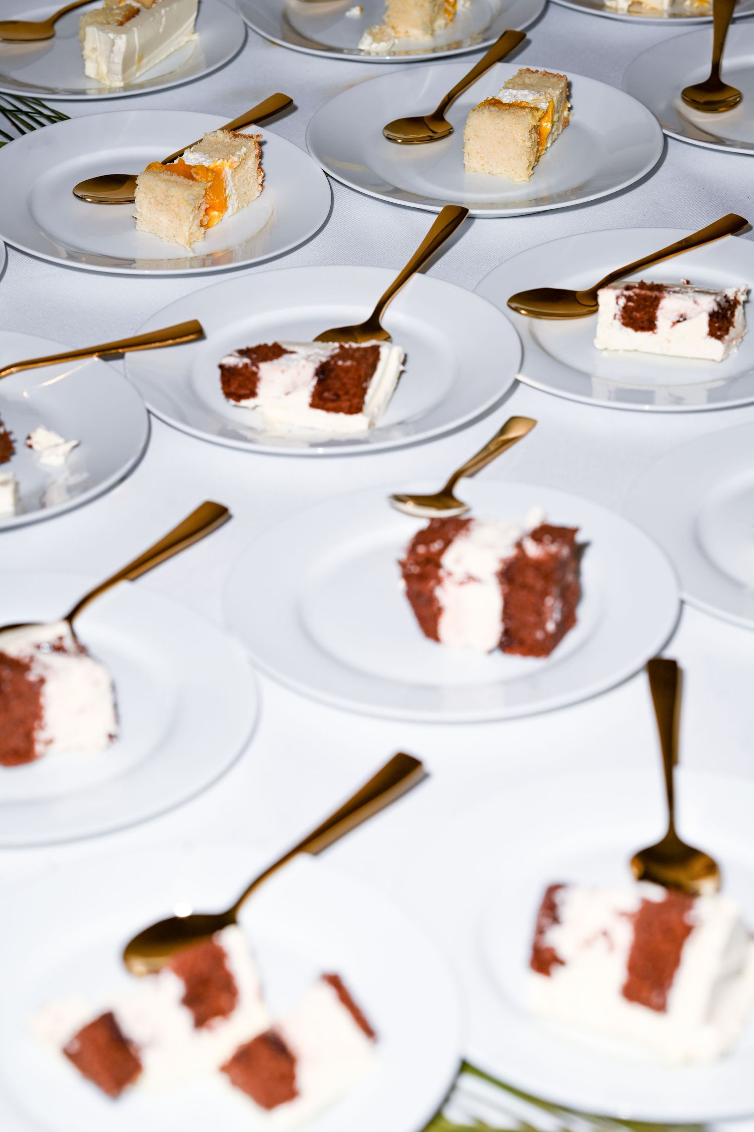 Multiple plates of wedding cake slices arranged with golden spoons on white tablecloth.