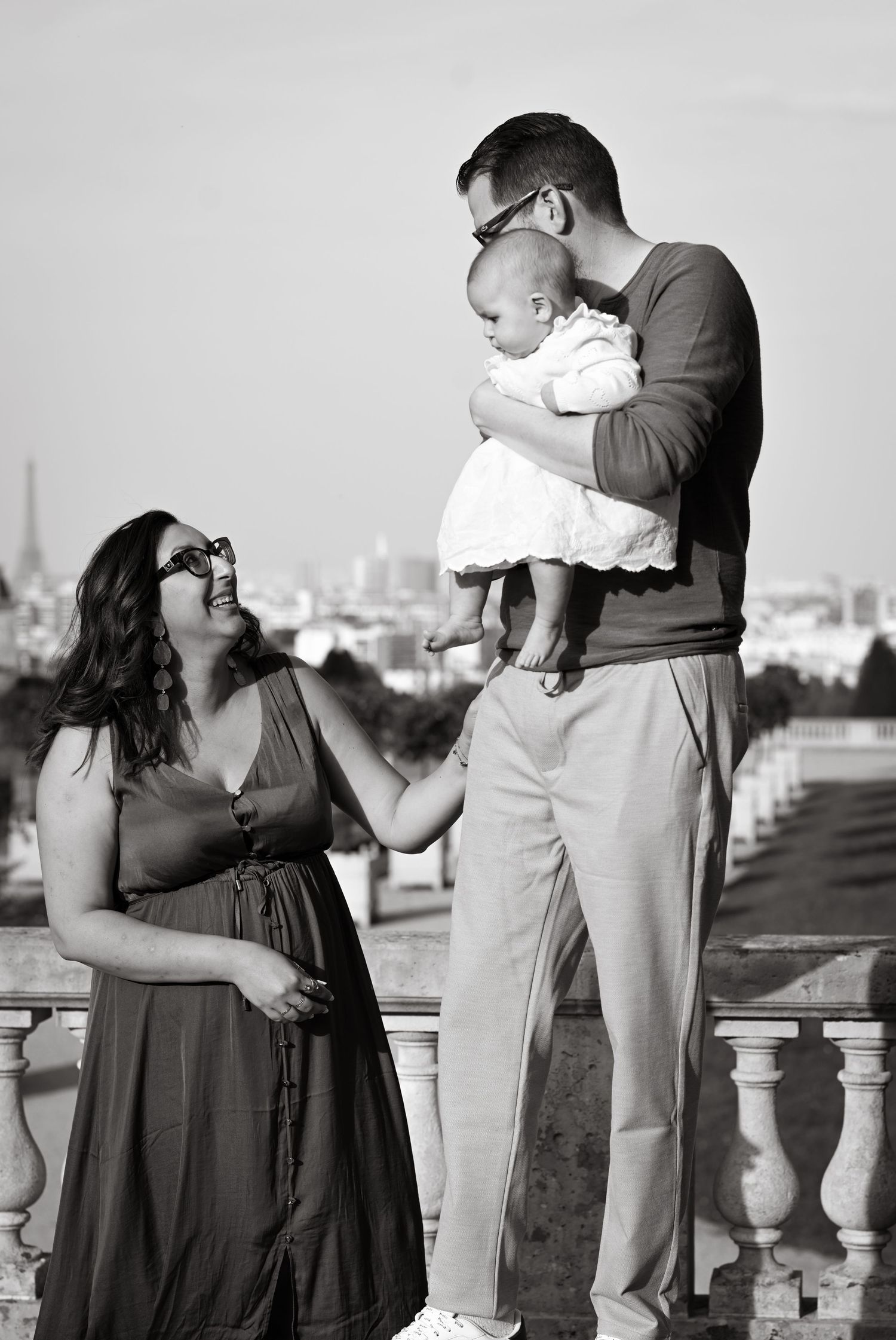 A family shares a sweet interaction on a balcony with a cityscape behind them.