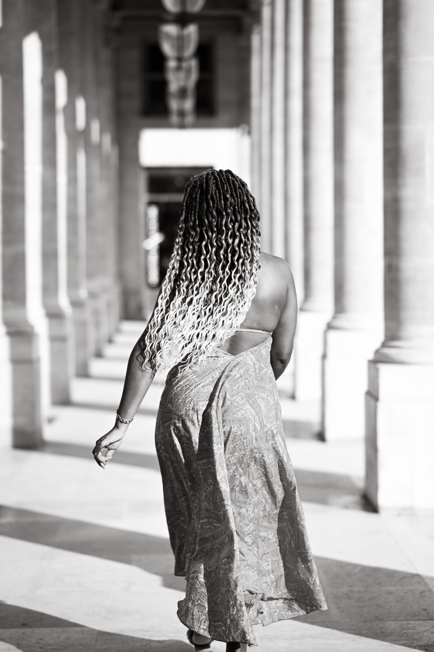Black and white portrait of long braided hair and flowing dress in columned hallway.