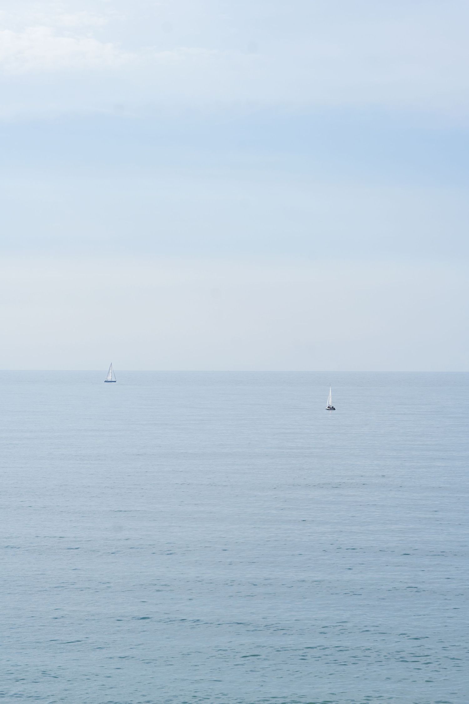Minimalist seascape with two small sailboats on calm waters under a pale blue sky.