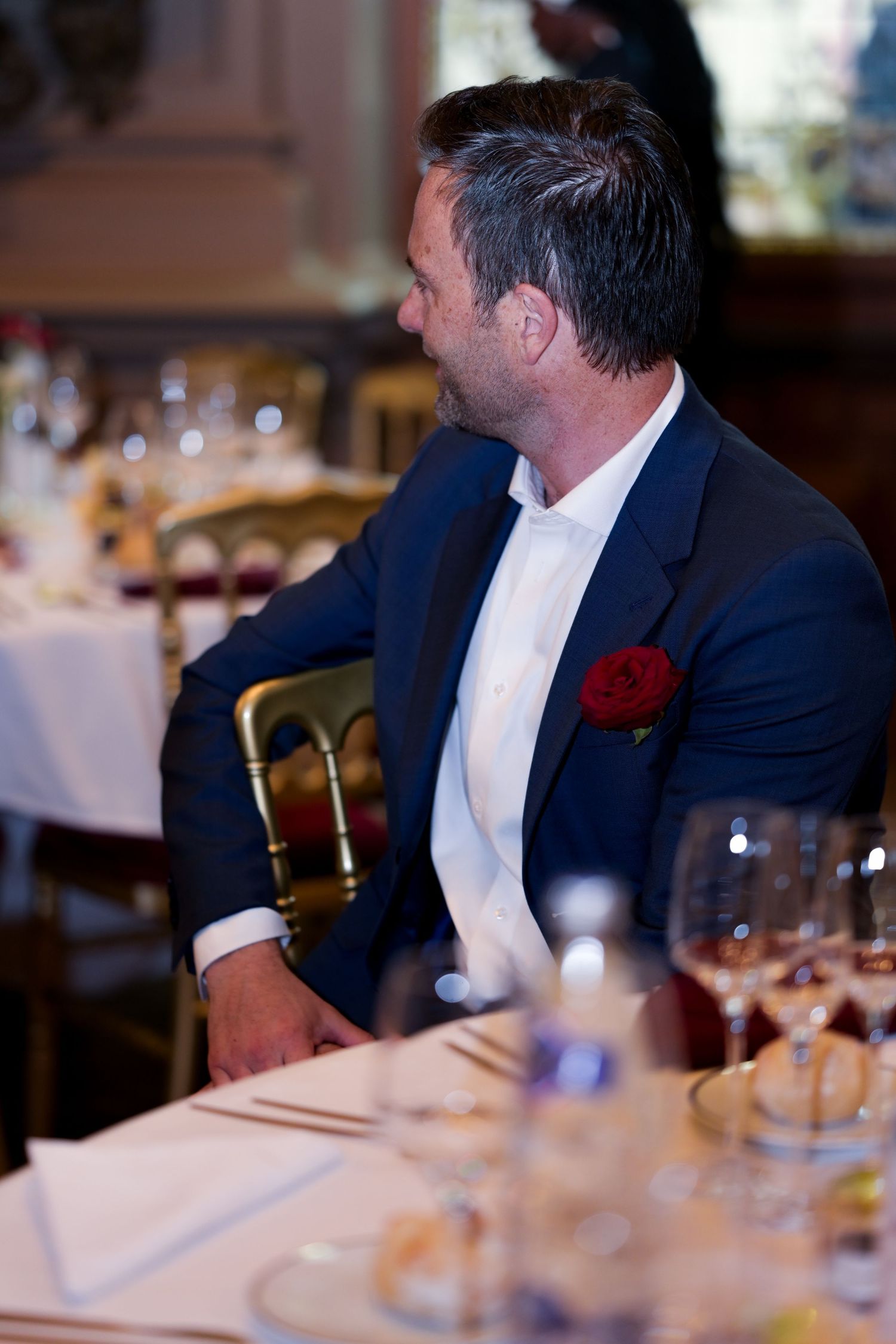 Guest in navy blazer and red pocket square seated at elegant dinner table.