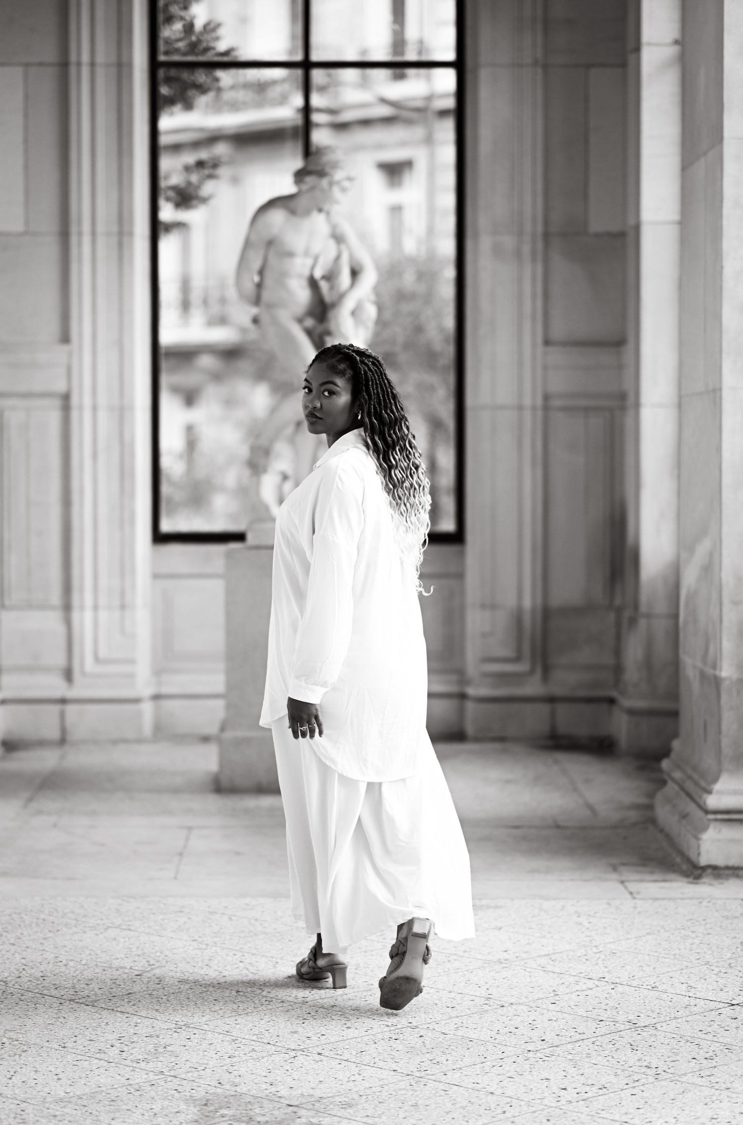 Figure in flowing white dress walks through elegant columned hallway in black and white photography.