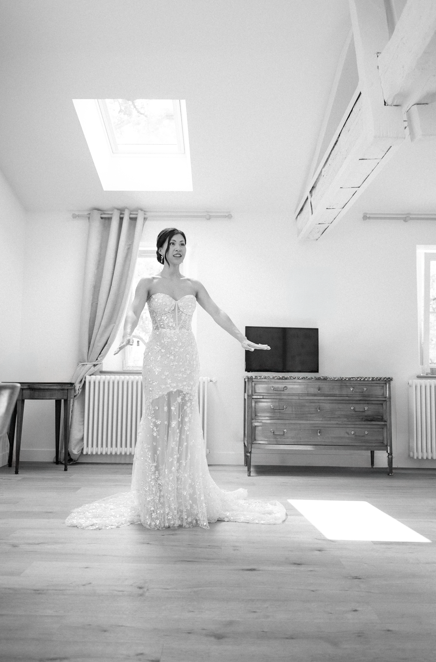 Bride in a strapless lace wedding gown with train poses in a sunlit room with hardwood floors and vintage dresser.