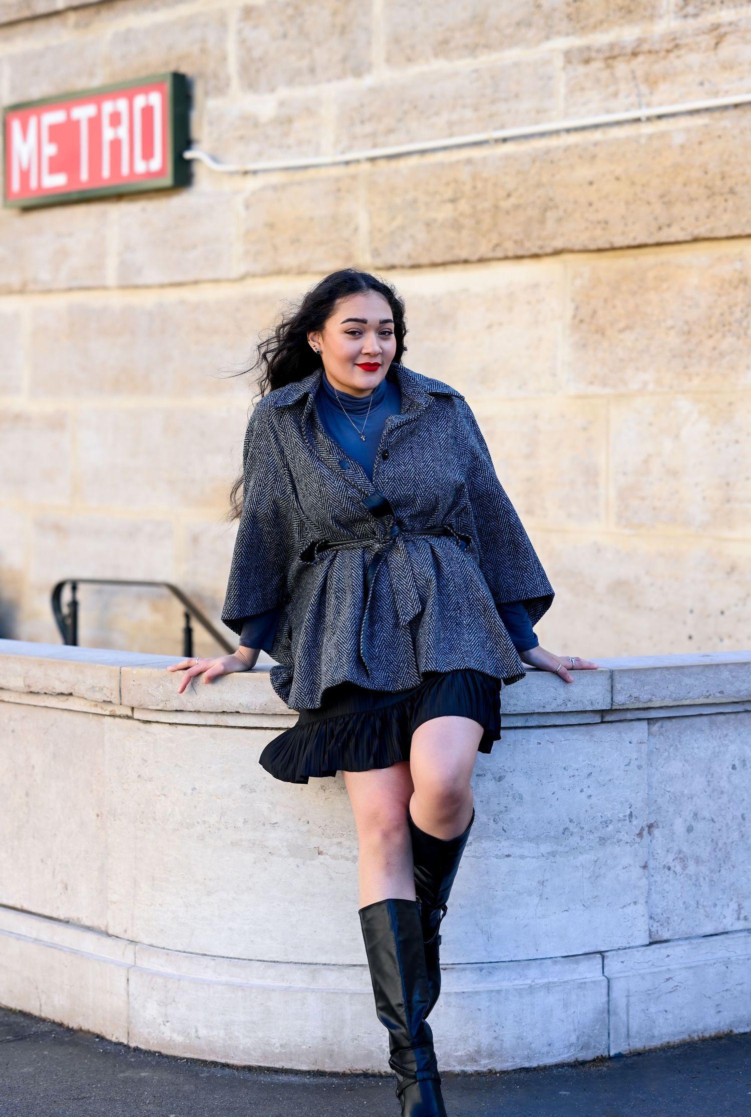 Stylish woman in belted grey coat and boots leans against stone wall near Metro entrance.