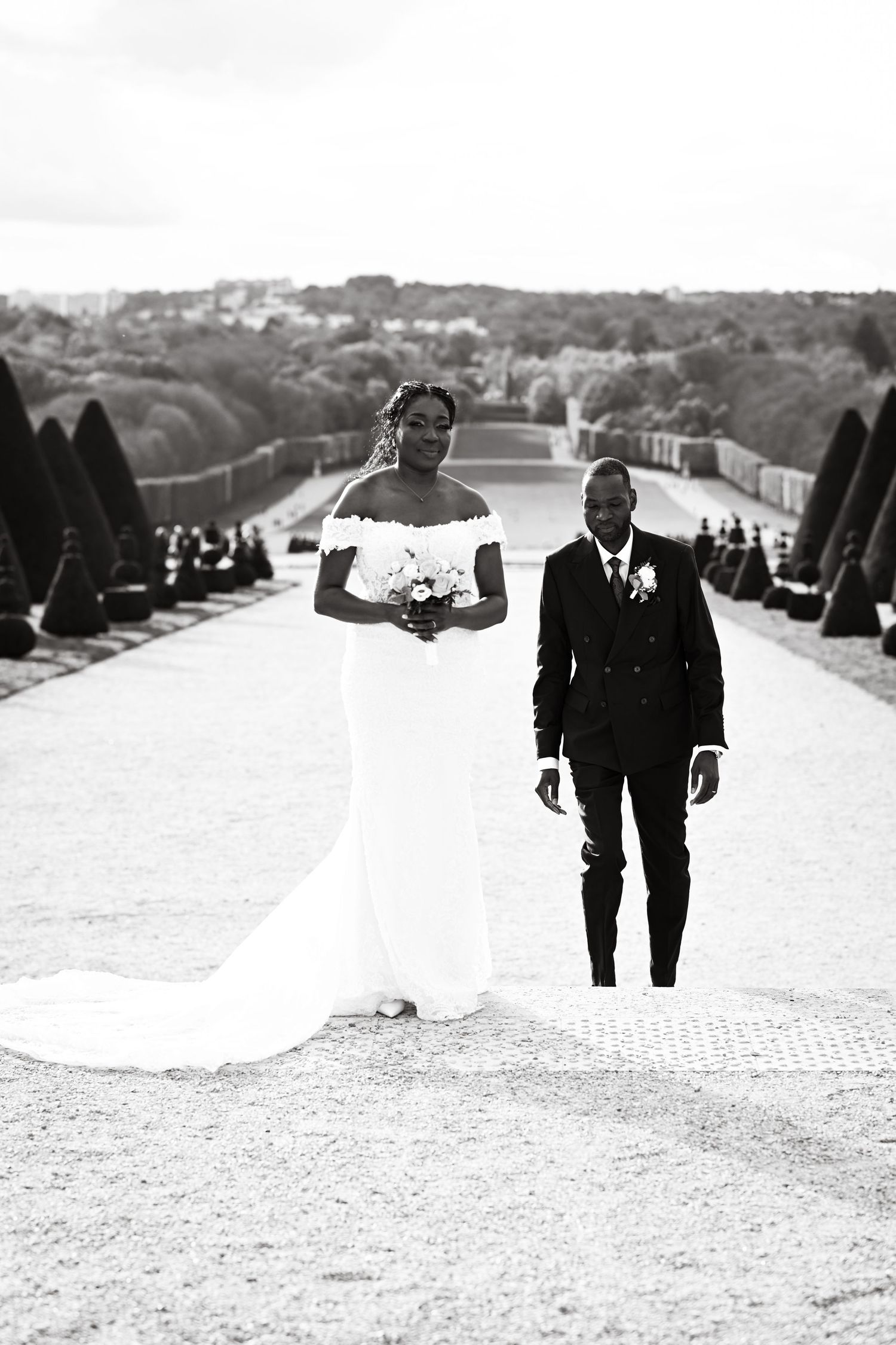 Black and white photo of newlyweds walking along a grand garden pathway with sculpted hedges.