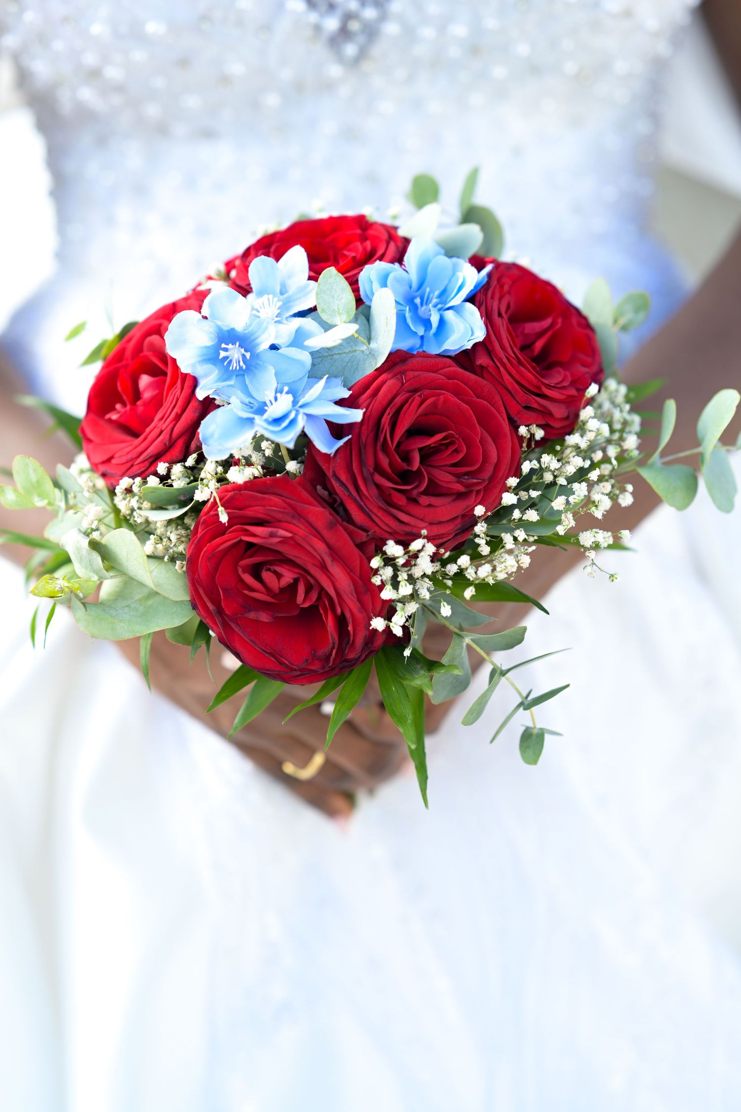 A wedding bouquet featuring red roses, blue flowers, and baby's breath against white fabric.