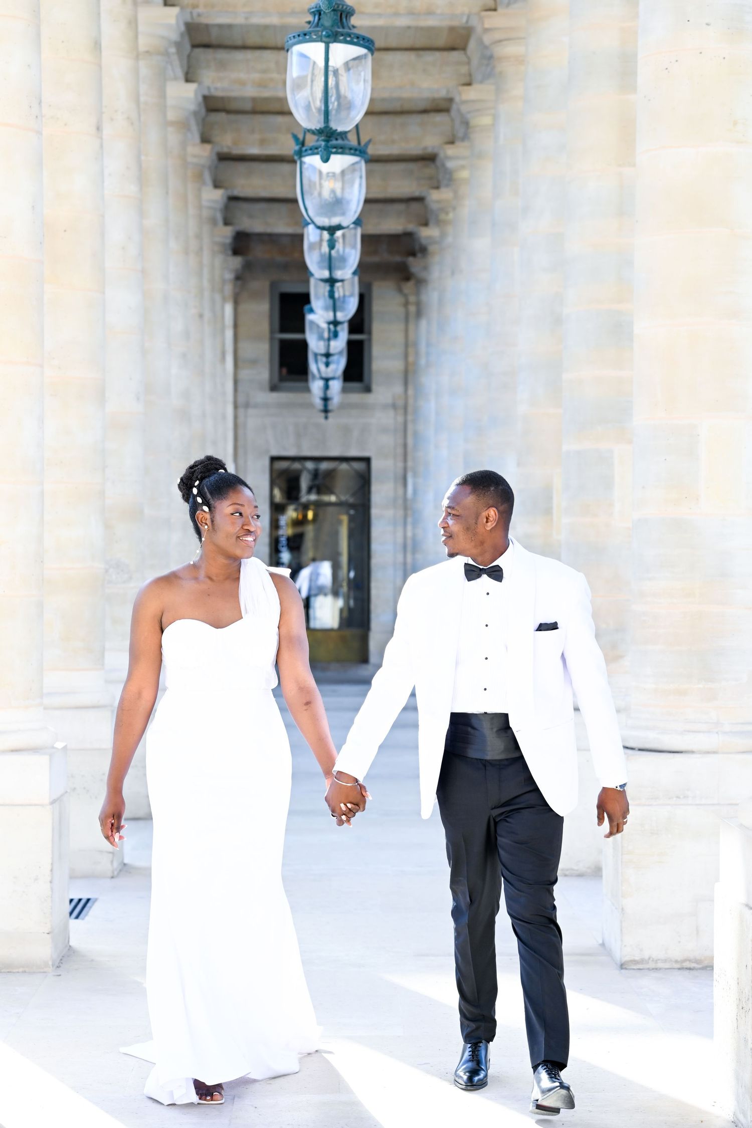 Newlyweds walking hand in hand through elegant columned hallway in formal attire.