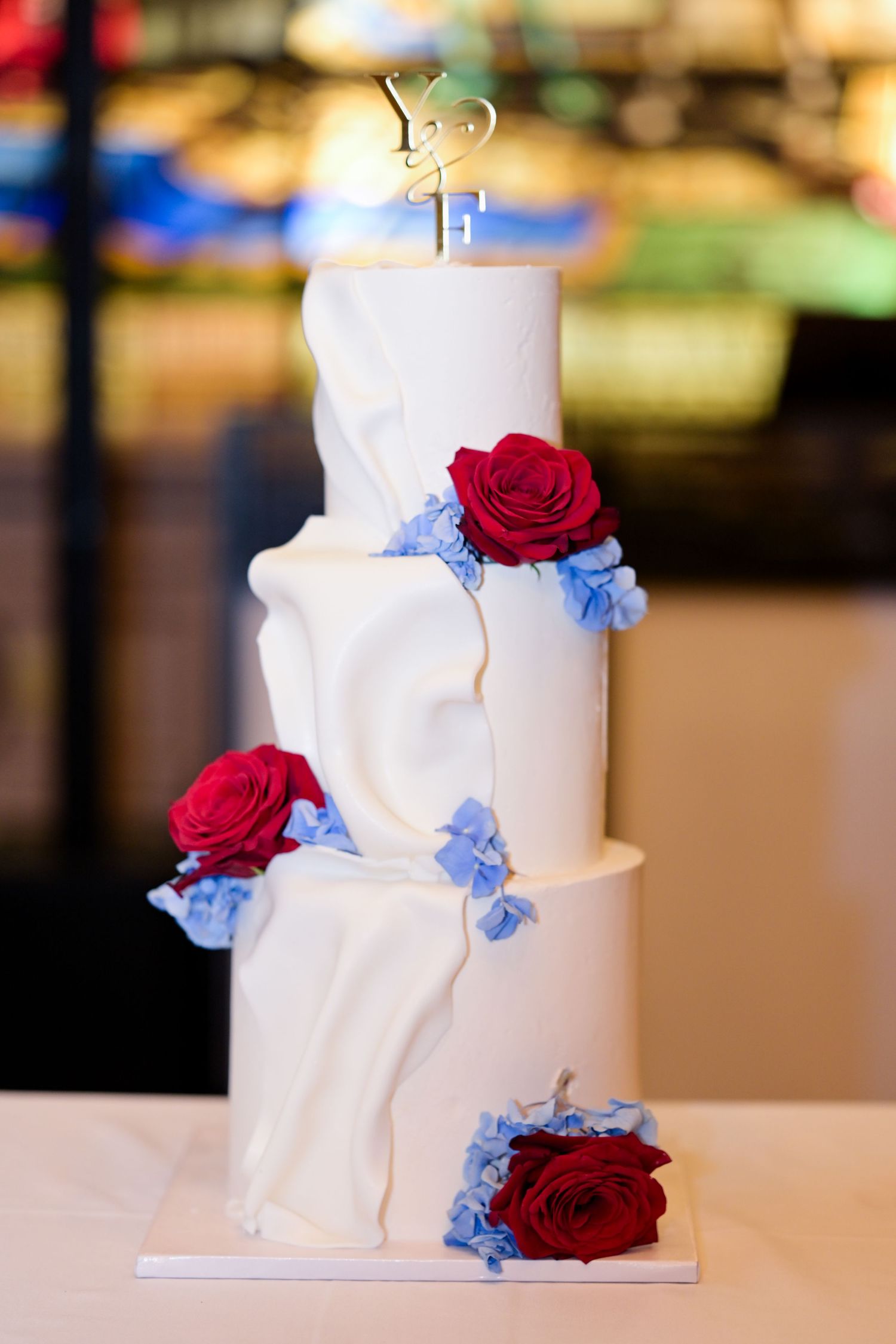 Elegant white wedding cake decorated with red roses and blue hydrangeas on a tiered display.