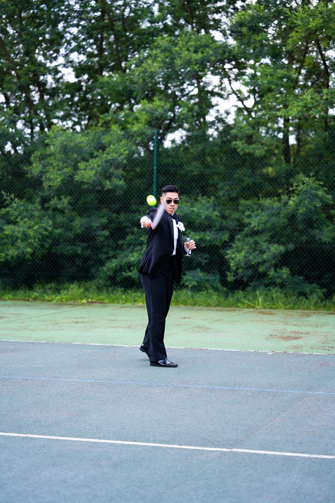 Person in tuxedo strikes dramatic pose on tennis court during wedding photoshoot.