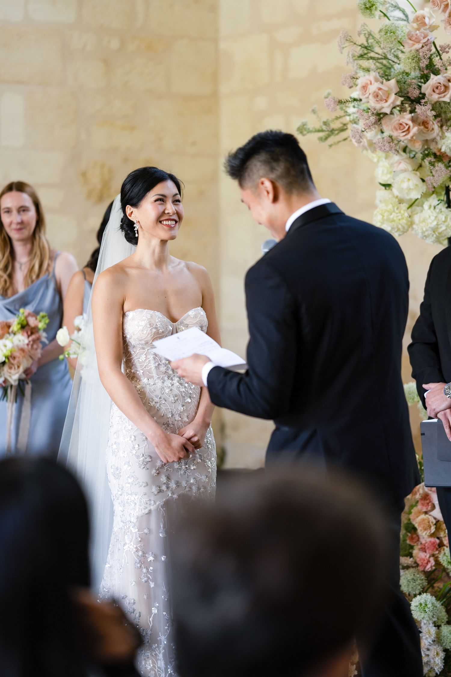 A wedding ceremony takes place with a couple exchanging vows under floral arrangements at an elegant indoor venue.