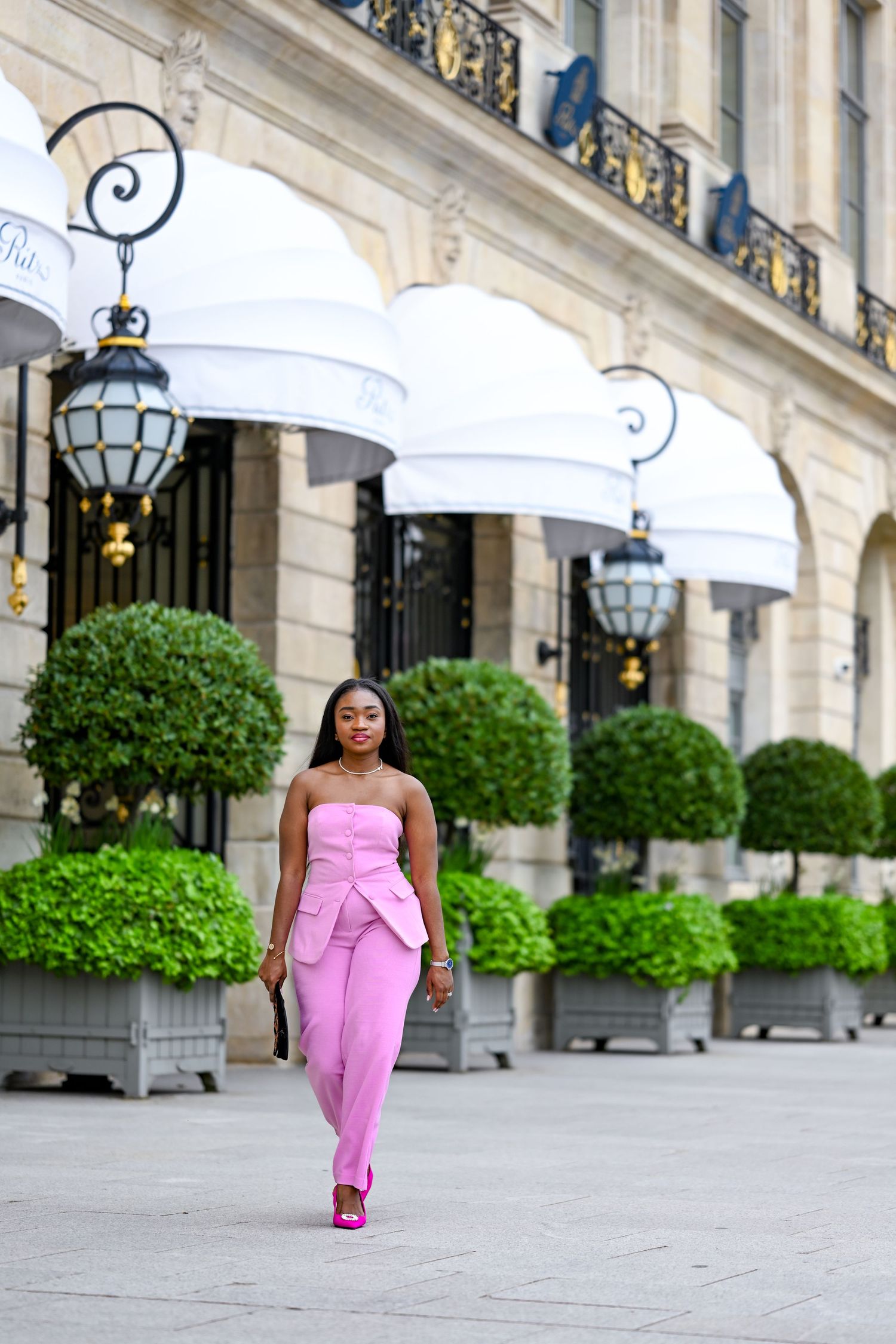 Woman in pink strapless jumpsuit walks confidently past luxury hotel facade with manicured topiaries.