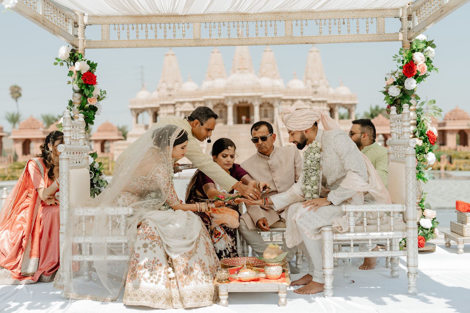 Outdoor wedding ceremony setup with white mandap, floral decorations and guests seated under blue sky.