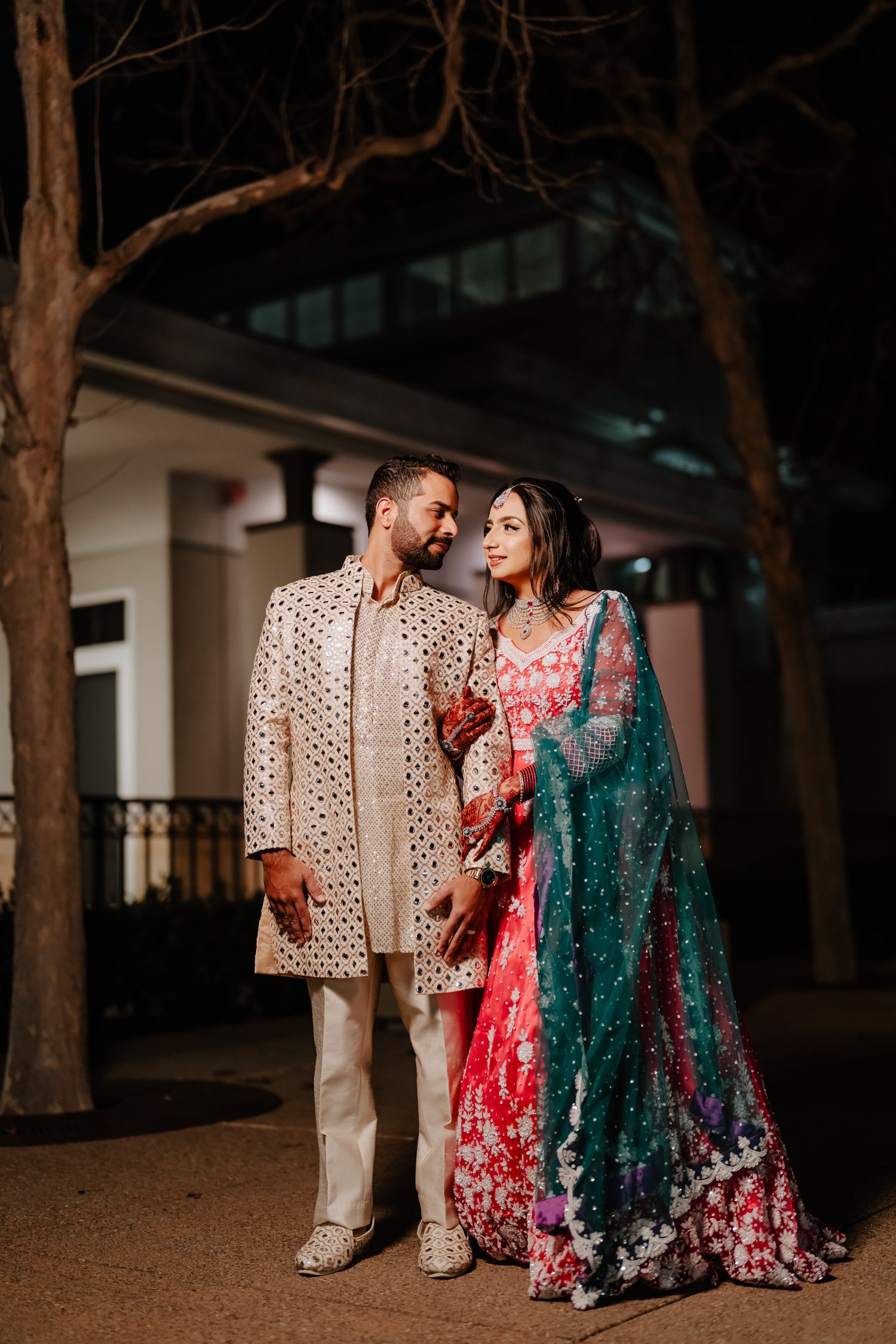 A couple in traditional South Asian wedding attire share romantic moments while walking outdoors at night.