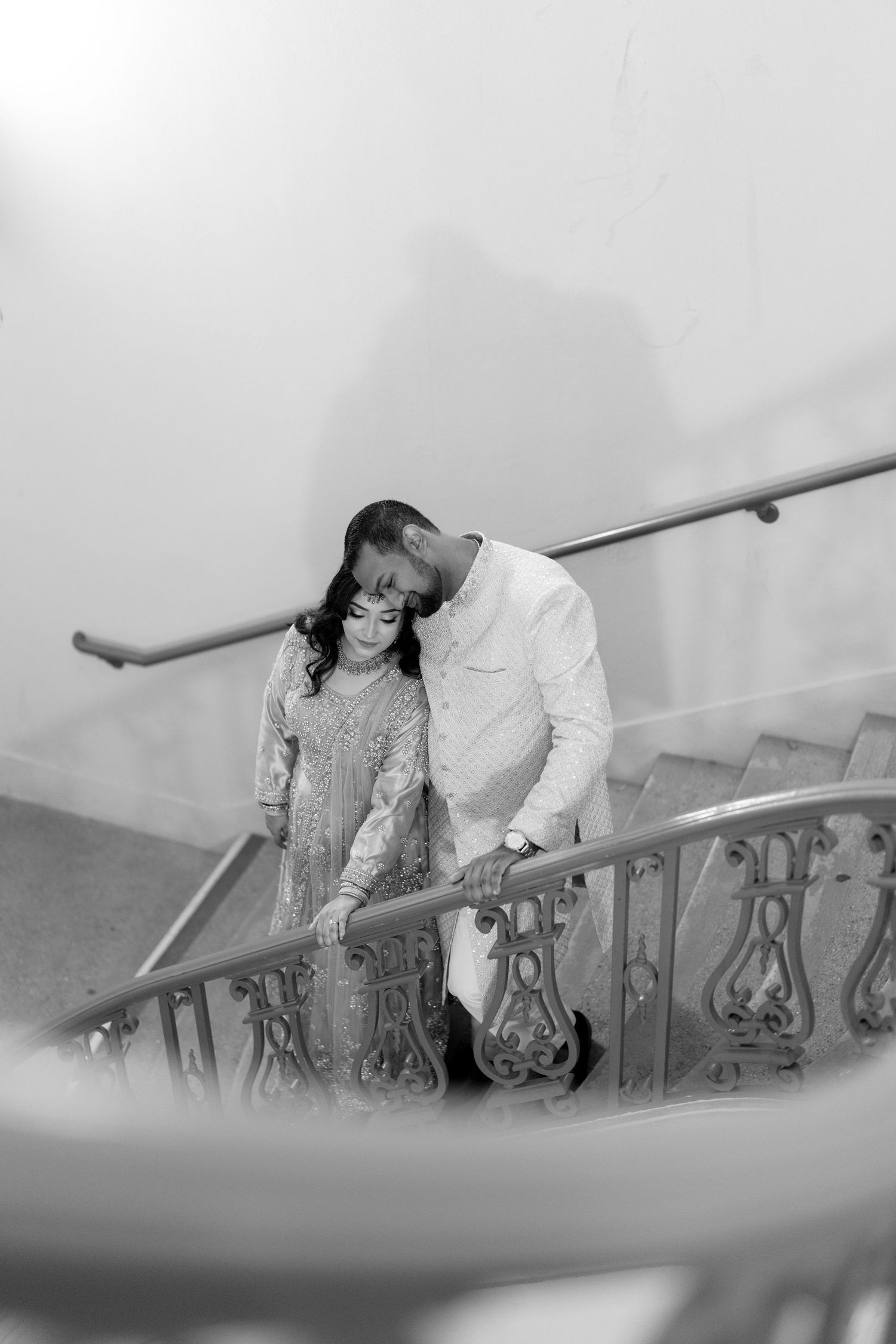 A couple in formal attire share a tender moment on an ornate staircase in black and white.