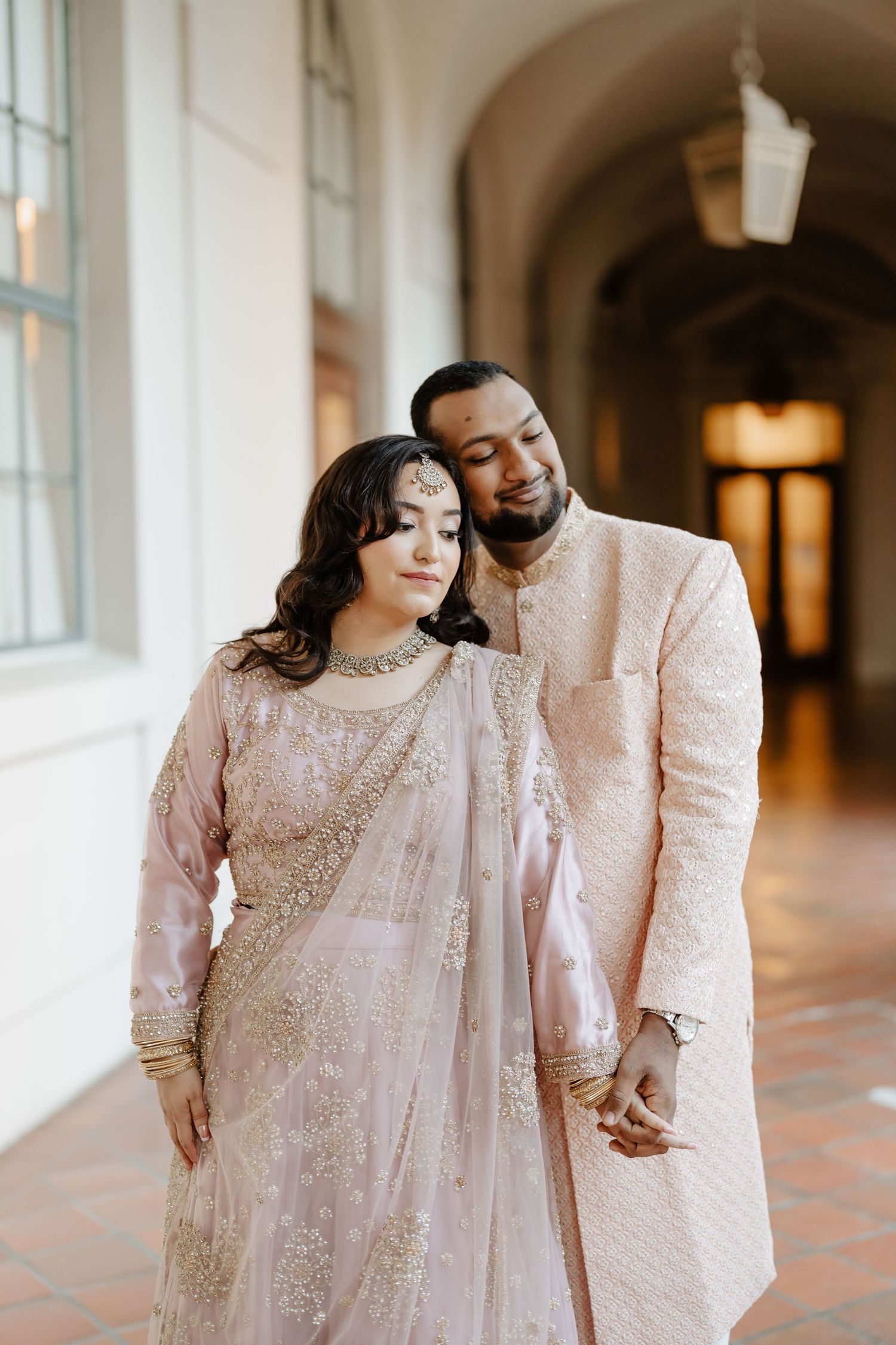 A couple in matching blush pink traditional South Asian wedding attire embrace in an elegant hallway.